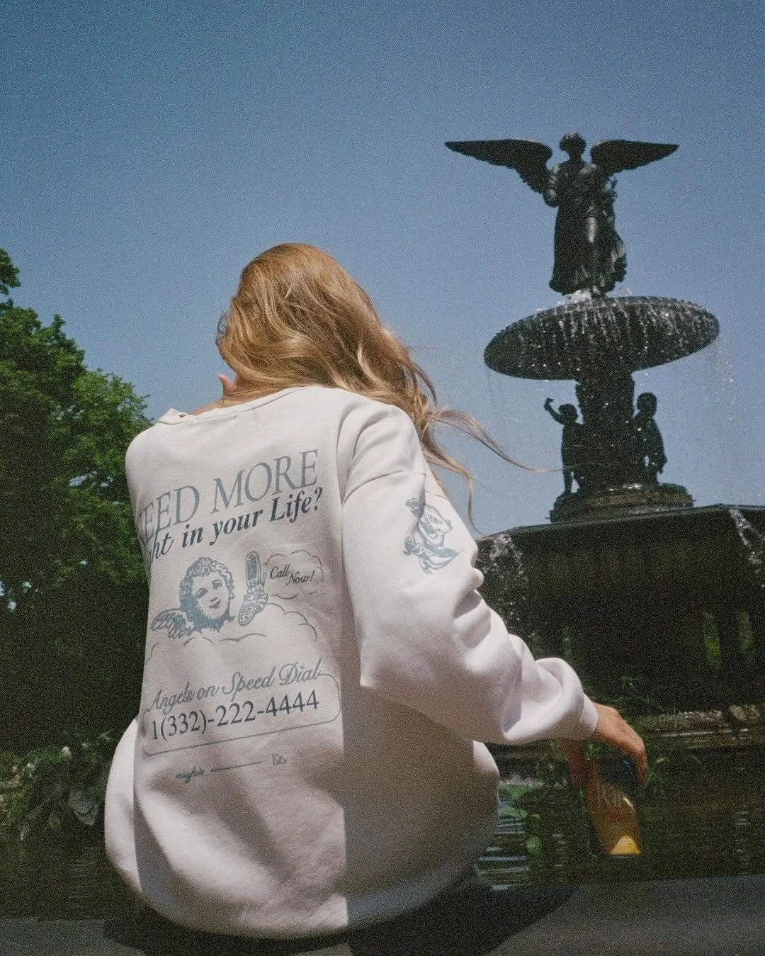 A woman with long hair wearing a white sweatshirt standing in front of a decorative water fountain with a sculpture of an angel with wings on top of the fountain, under a clear blue sky.