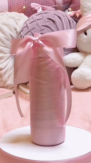 A tall pink decorative vase wrapped with a pink ribbon bow, placed on a white surface in a cozy background with plush pillows and stuffed animals.