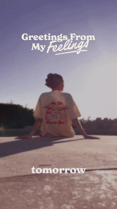 Person sitting on a concrete surface outdoors during sunset, facing away from camera, with text overlay saying "Greetings From My Feelings" and "tomorrow".
