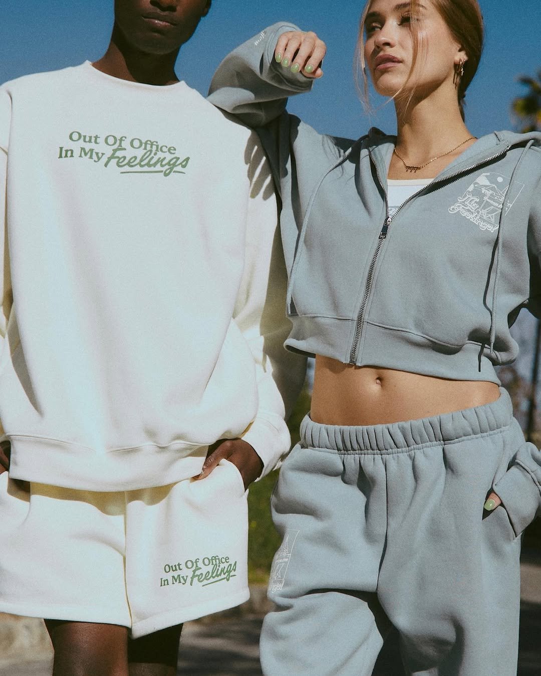 Two women wearing casual athletic outfits outdoors, with one in a white sweatshirt and matching shorts, and the other in a gray cropped hoodie and sweatpants. The sweatshirts have the phrase 'Out Of Office In My Feelings' printed on them.