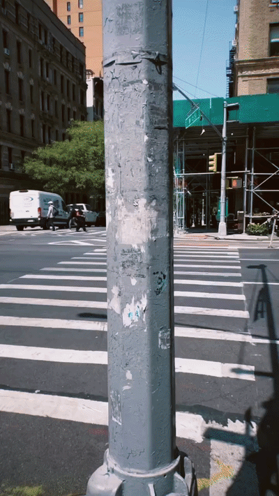 A weathered metal pole in front of a city crosswalk, with buildings and scaffolding in the background on a sunny day.