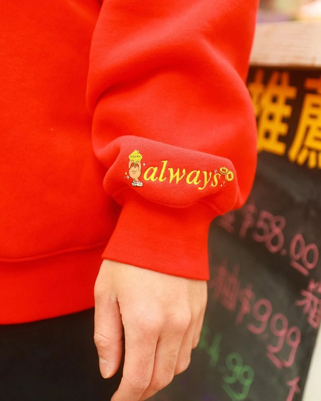 Close-up of a red sweatshirt sleeve with the word 'always' embroidered in yellow, accompanied by a small cartoon hand making a thumbs-up gesture with the word 'LUCKY' on it. Part of a colorful chalkboard with writing is visible in the background.
