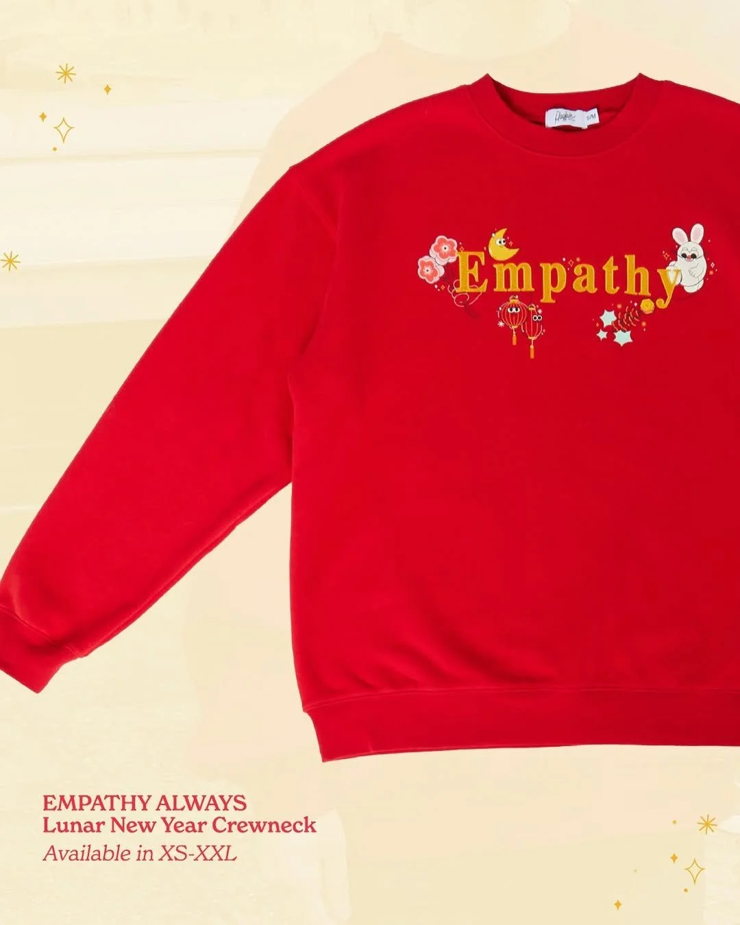 A red crewneck sweatshirt with the word "Empathy" printed in yellow, surrounded by small illustrations of flowers, a rabbit, a moon, lanterns, and stars, with additional text advertising it as part of the Lunar New Year collection available in sizes XS to XXL.