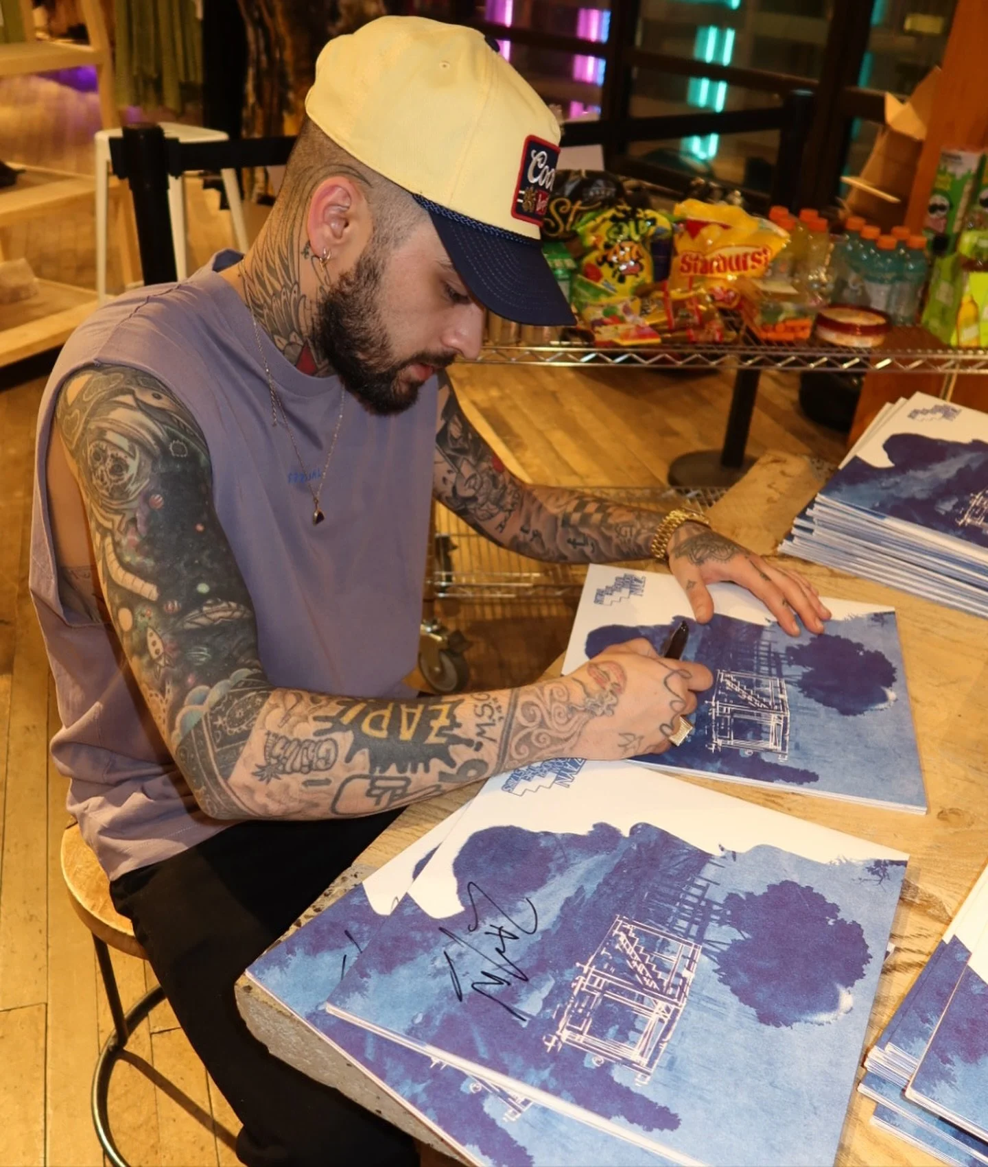 A tattooed man wearing a beige cap writes on blue and white posters at a table in a store or event space with shelves of snacks and drinks in the background.