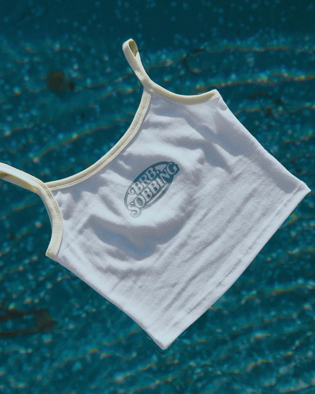 A white sleeveless shirt with a logo that reads 'Goblin Sobbin' in a spiral, hanging in mid-air against a textured blue background.