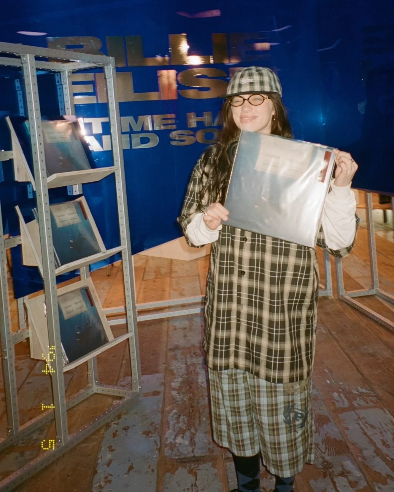 A woman smiling and holding a vinyl record in a store with metal display racks containing more records, against a blue wall with partial text.