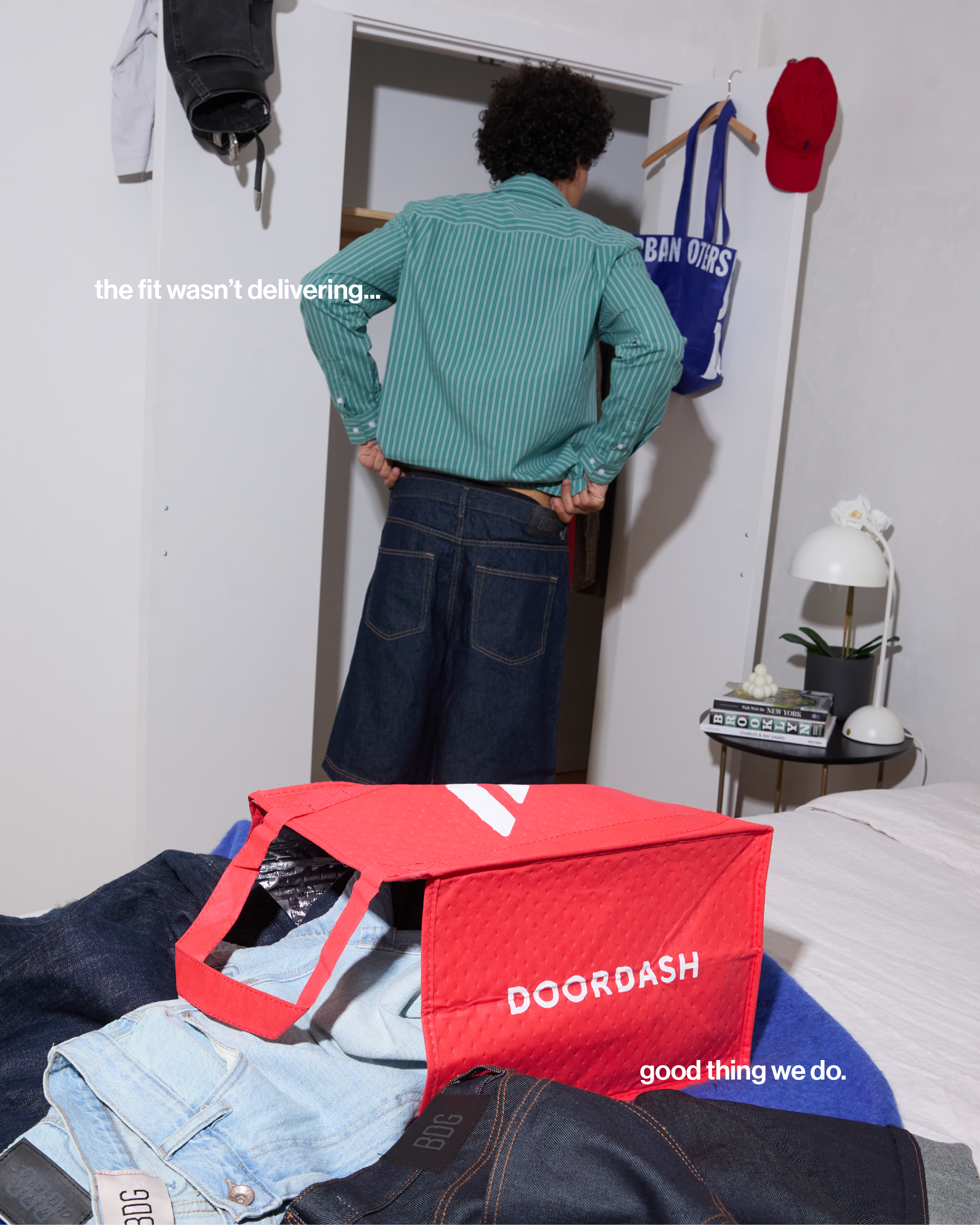 A person with curly hair looking into an open closet door. There are hats and bags hung on the door. The person appears to be organizing or choosing clothes. On the bed, there's a red Doordash delivery bag containing clothing items. The room has a small black side table with a lamp, a plant, and some magazines. The overall scene depicts someone sorting laundry or packages in a bedroom.