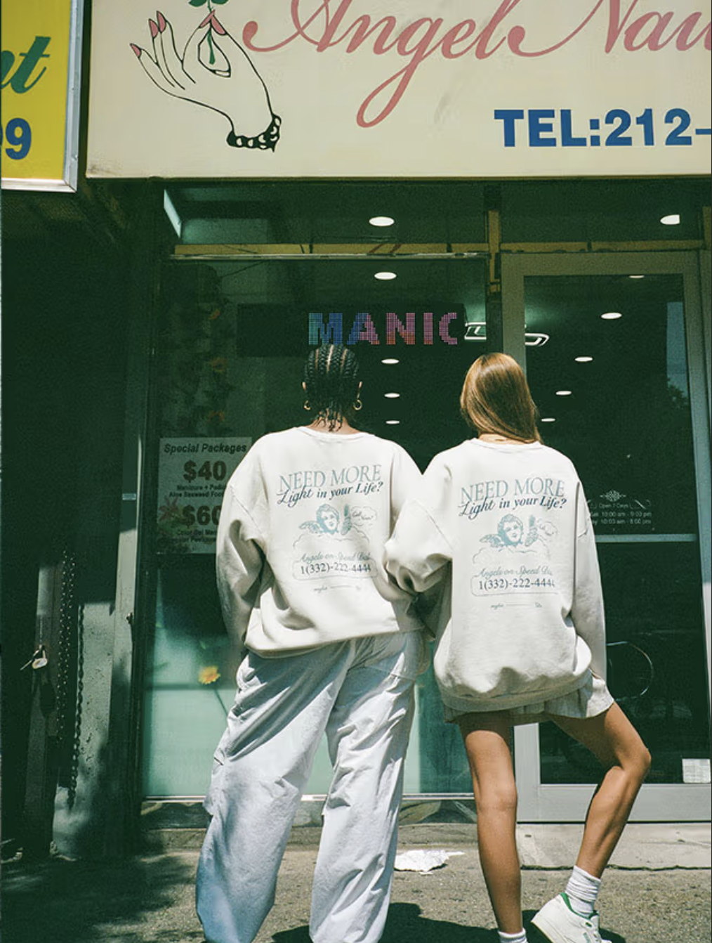 Two women wearing matching sweatshirts with the phrase "Need More Light in your Life?" on the back, standing in front of a storefront with a sign that reads "Angel Nails" and tinted glass windows.