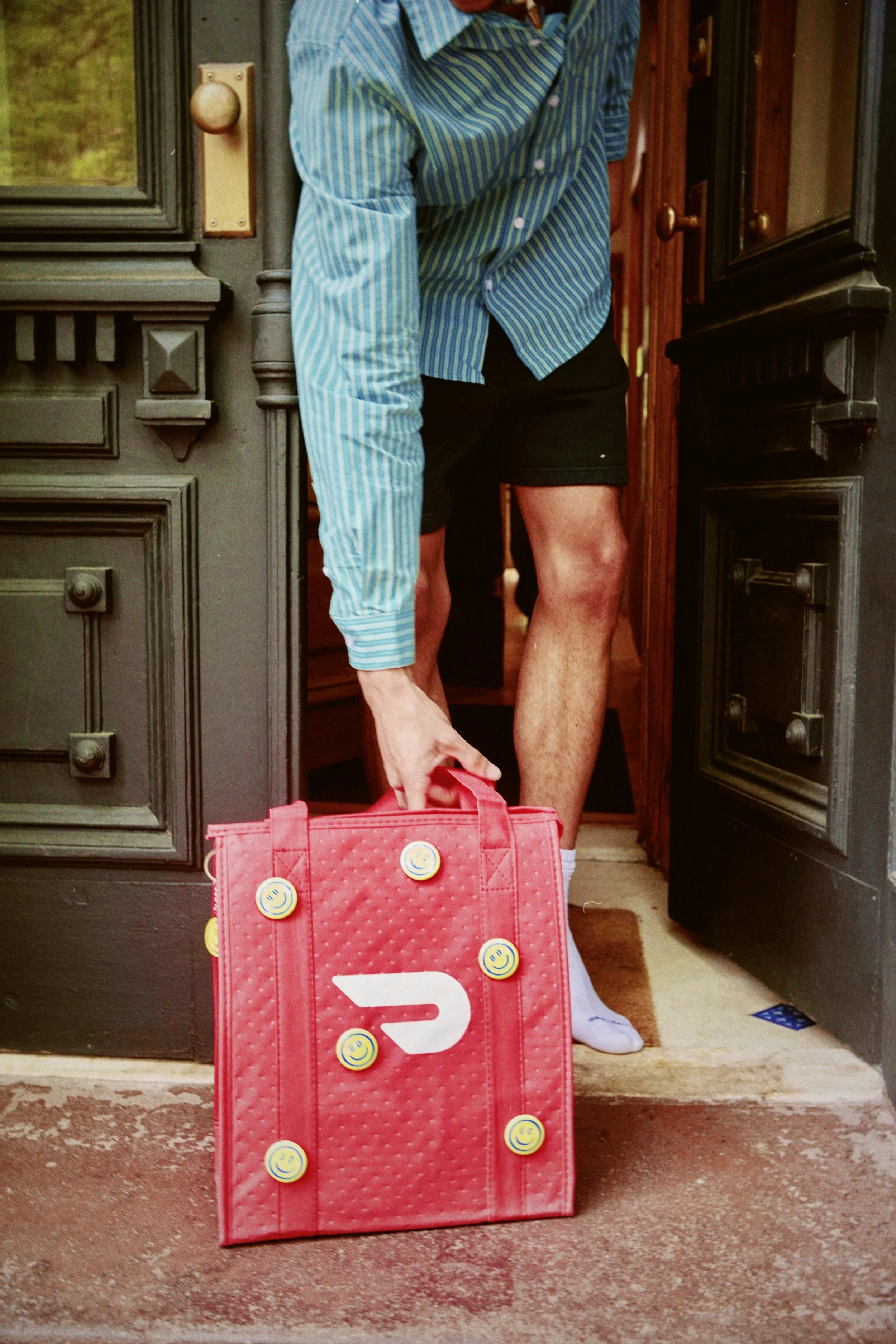 Person wearing a blue striped shirt and black shorts stepping through a doorway, holding a pink bag with smiley face stickers in front of a dark door.