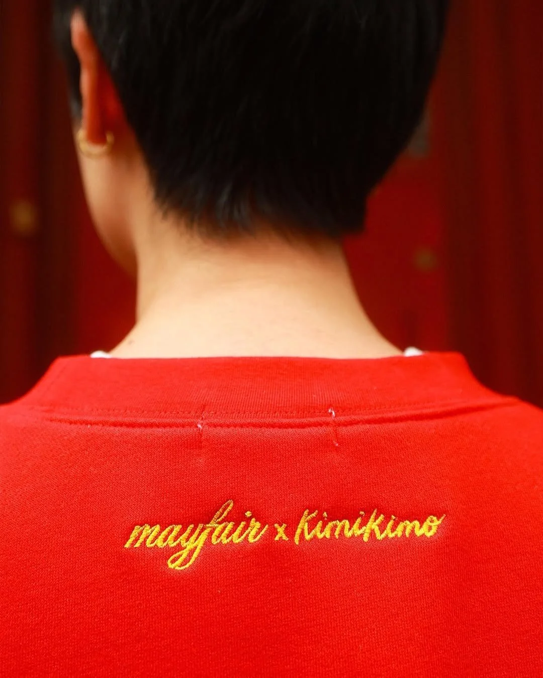 Back of a person wearing a red shirt with gold embroidery that says 'mayfair x kimkkimo'