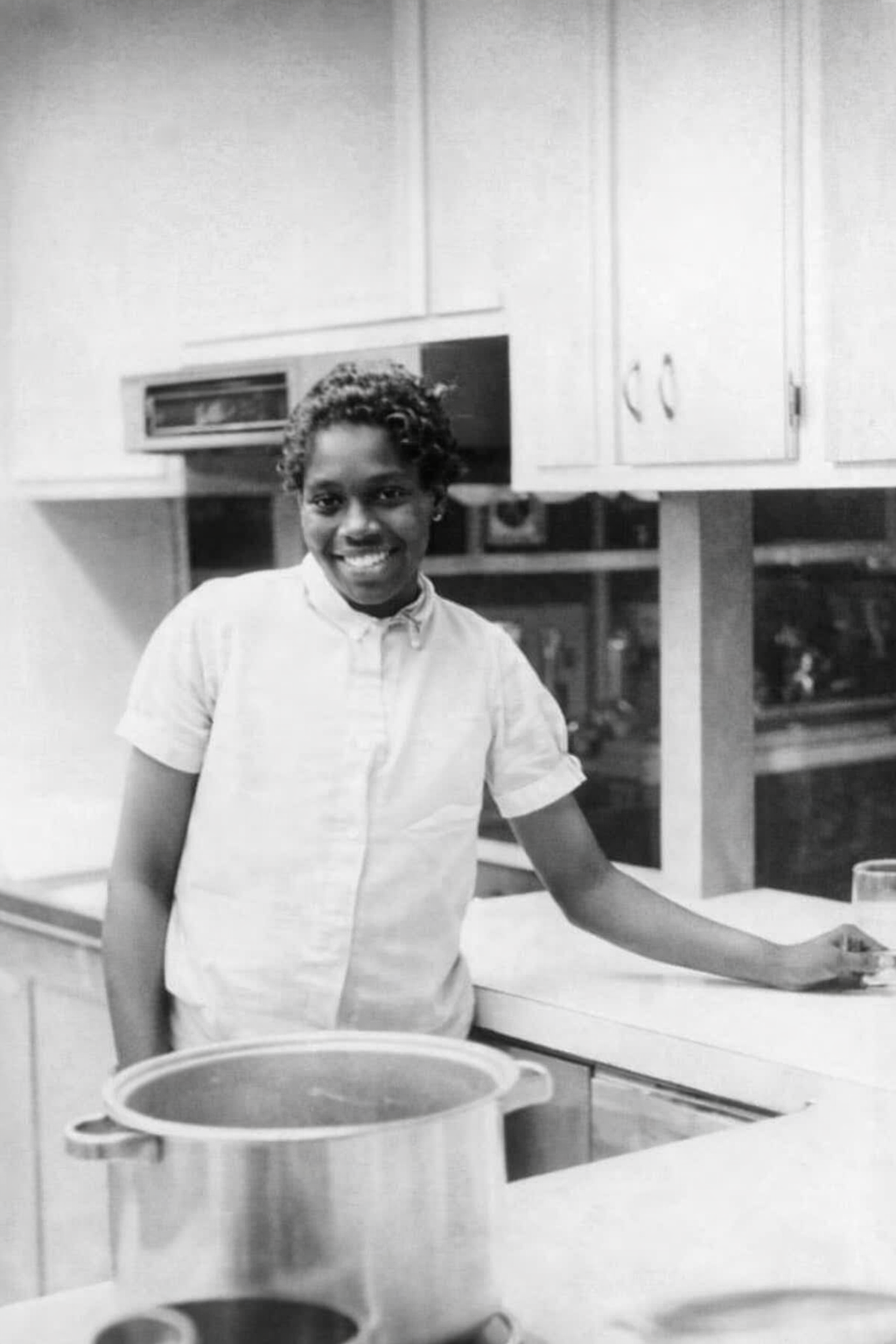 A woman with short curly hair and dark skin smiling while standing in a kitchen near a counter, wearing a light-colored short-sleeved shirt. There are cabinets above and a pot on the counter in front of her.