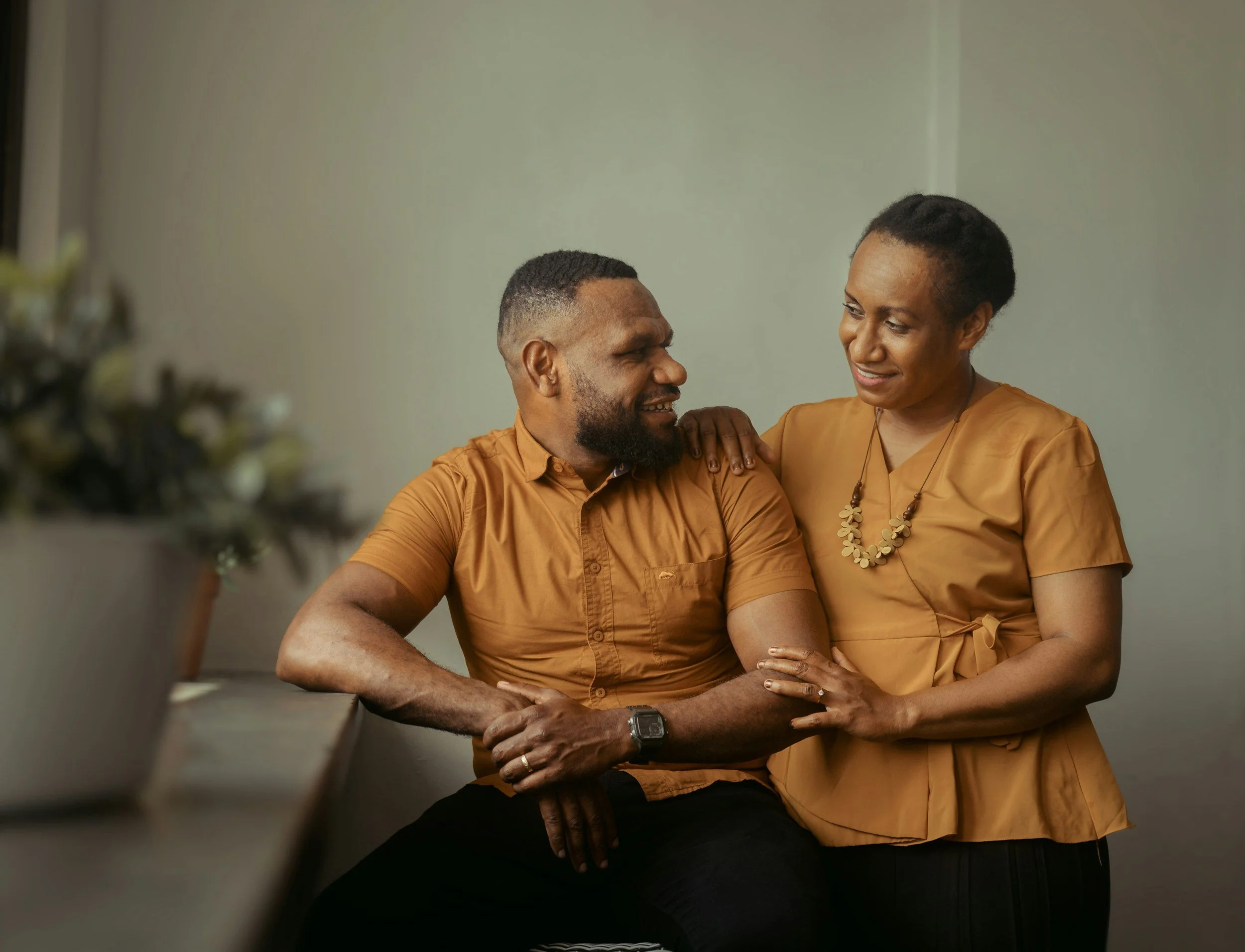 A man and woman sharing an intimate moment indoors, smiling at each other. The woman is wearing an orange dress with a bead necklace, and the man is wearing a similar orange shirt with a watch on his wrist.