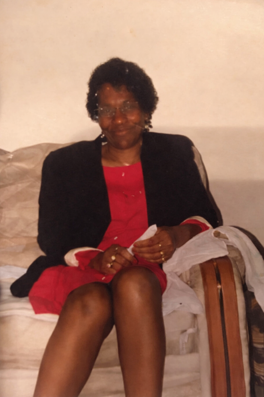 A woman with short curly hair and glasses sitting on a sofa, wearing a black jacket over a red dress, with her legs crossed.