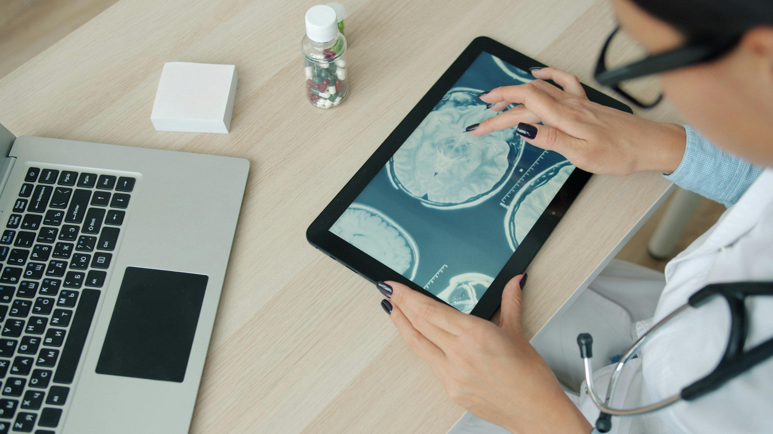 A doctor using a tablet with brain MRI images displayed on the screen, on a light wooden desk with a laptop, medicine bottle, and a white box.