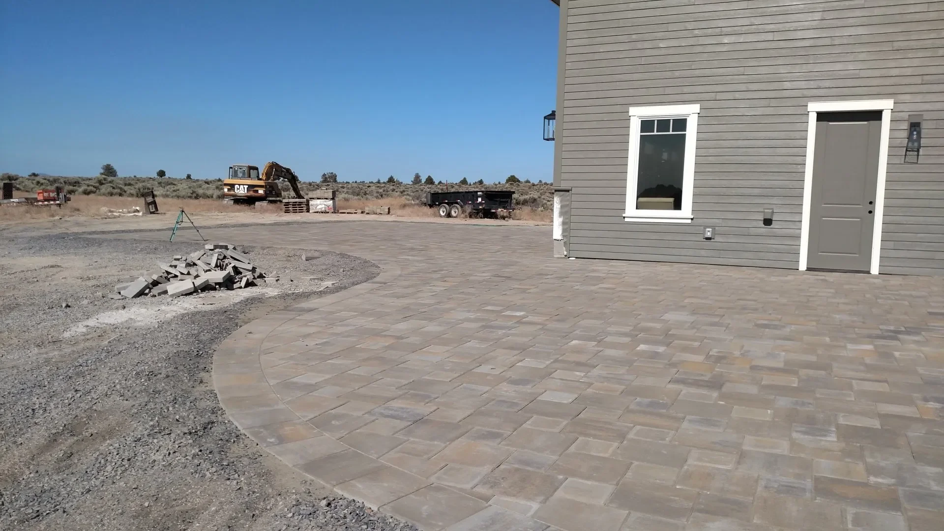 Construction site with a partially paved outdoor area, a gray house with white trim, a window, and a door, with construction materials and equipment, including a yellow excavator and a trailer, in the background under a clear blue sky.