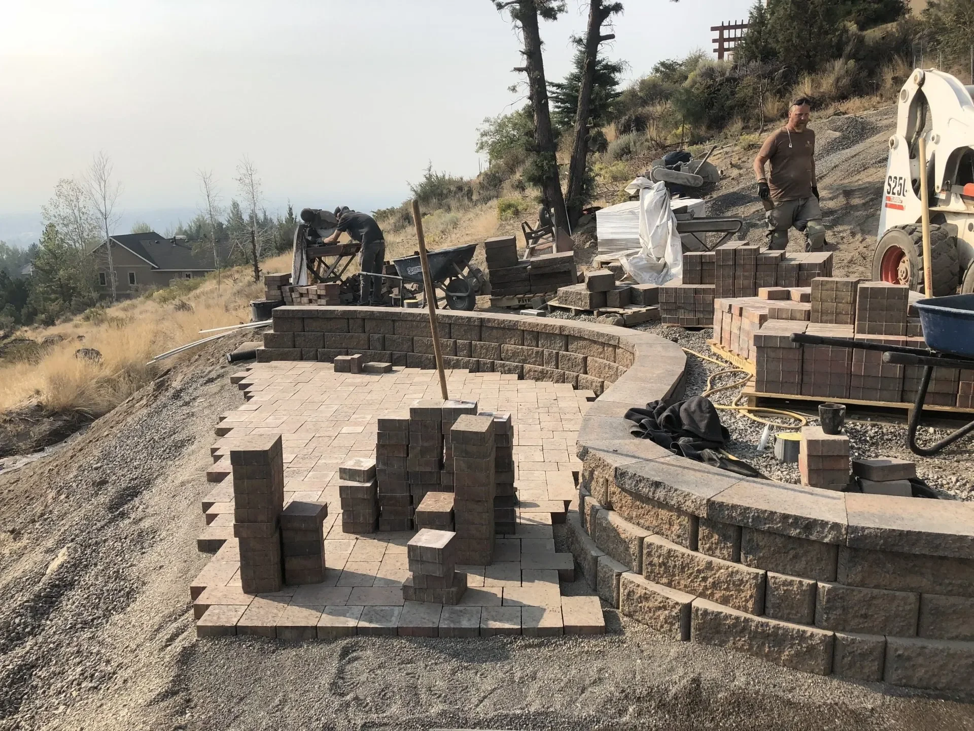 Workers constructing a stone patio on a hillside using bricks and pavers, with trees and a house in the background.