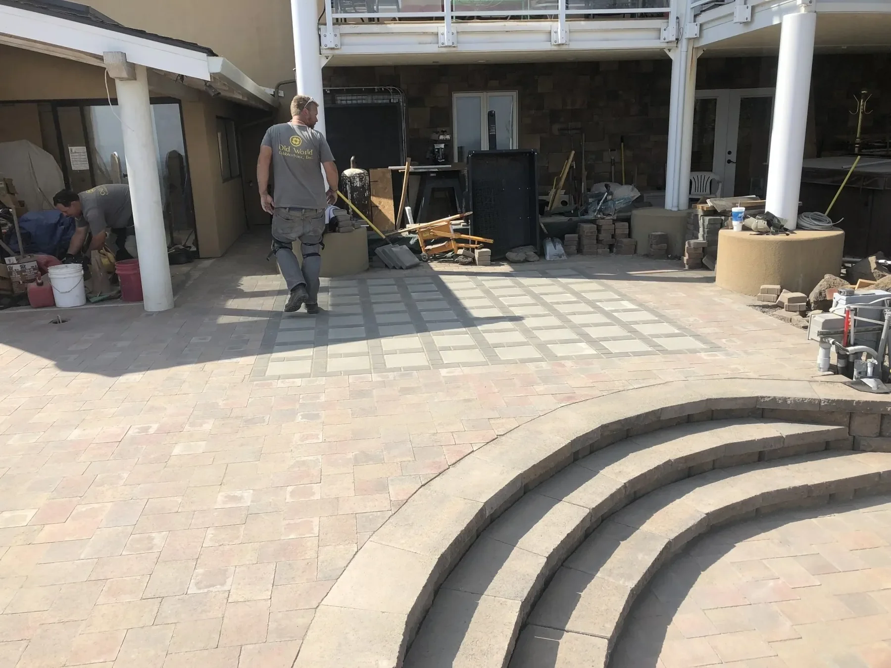 Workers installing interlocking pavers on a backyard patio area, with construction tools and materials visible. Paved area has a shaping pattern, and there are stone steps in the foreground.
