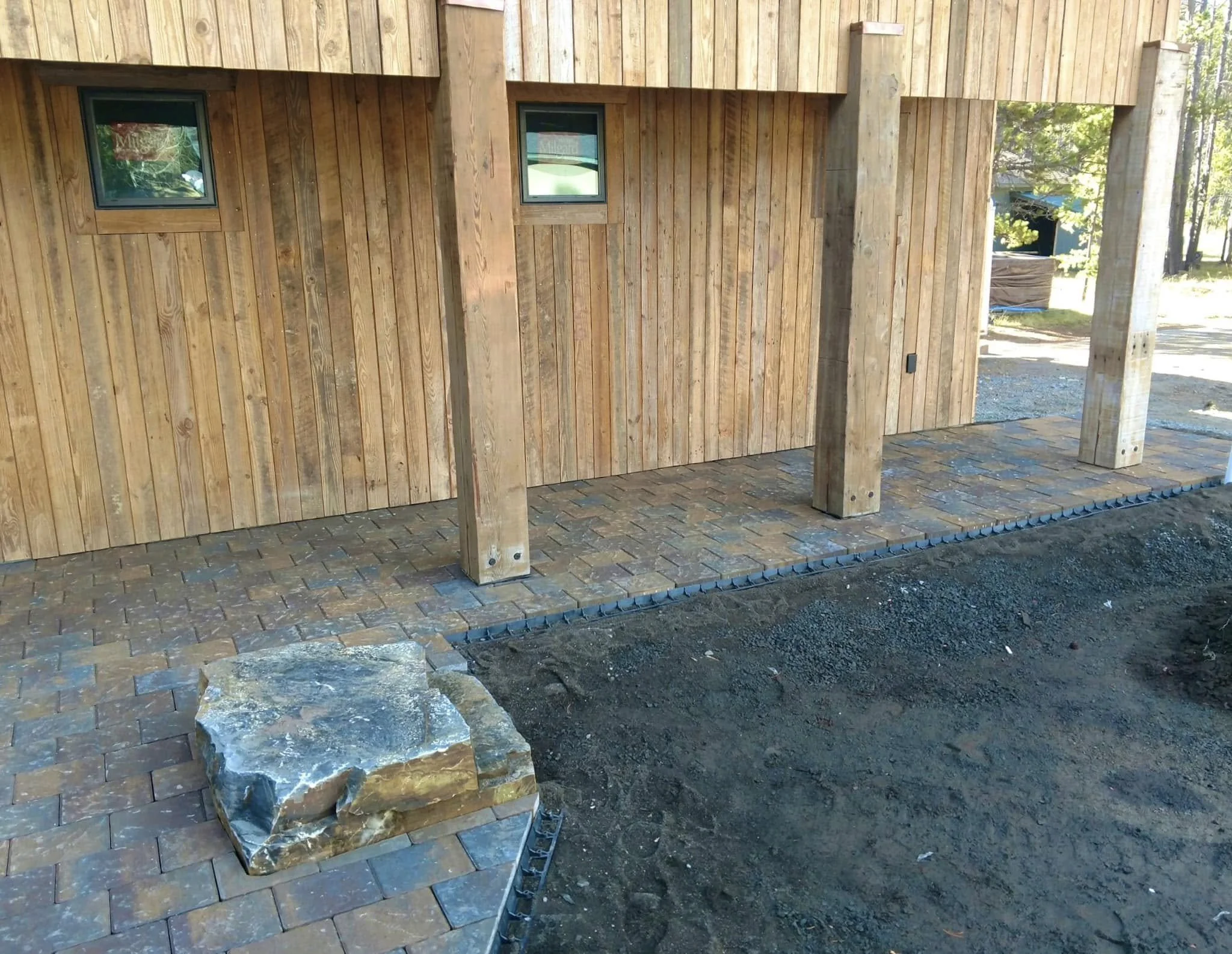 Back side of a wooden house under construction with window and brick patio, surrounded by trees.