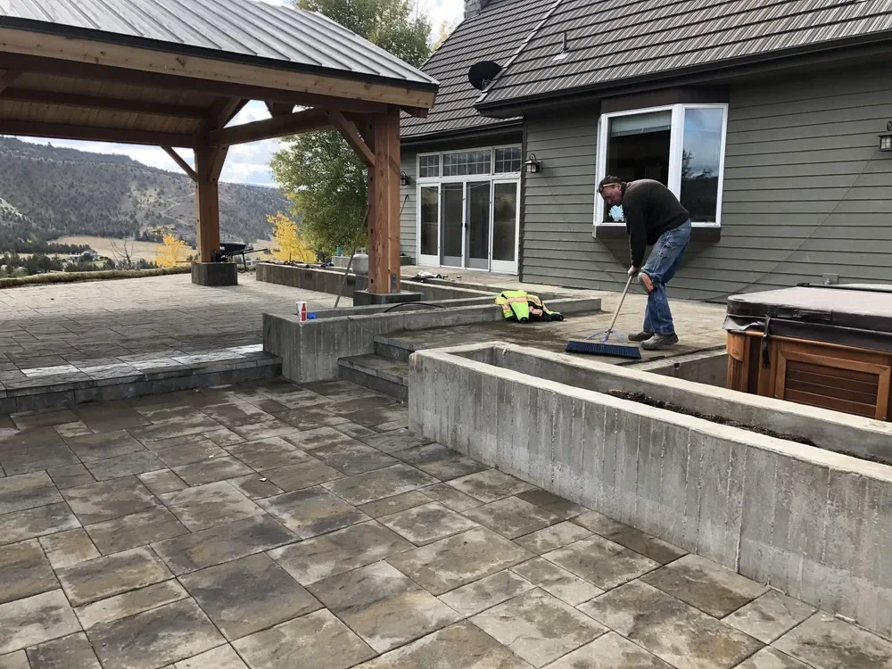 A person sweeping a concrete patio outside a house with gray siding and sliding glass doors. The patio has stone tiles and an outdoor hot tub nearby, with hills and trees in the background.