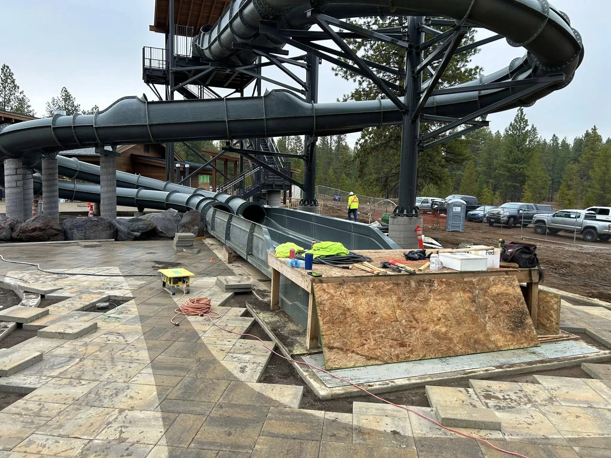 Construction site with a large water slide structure, workers in safety vests, and paving stone work in progress.