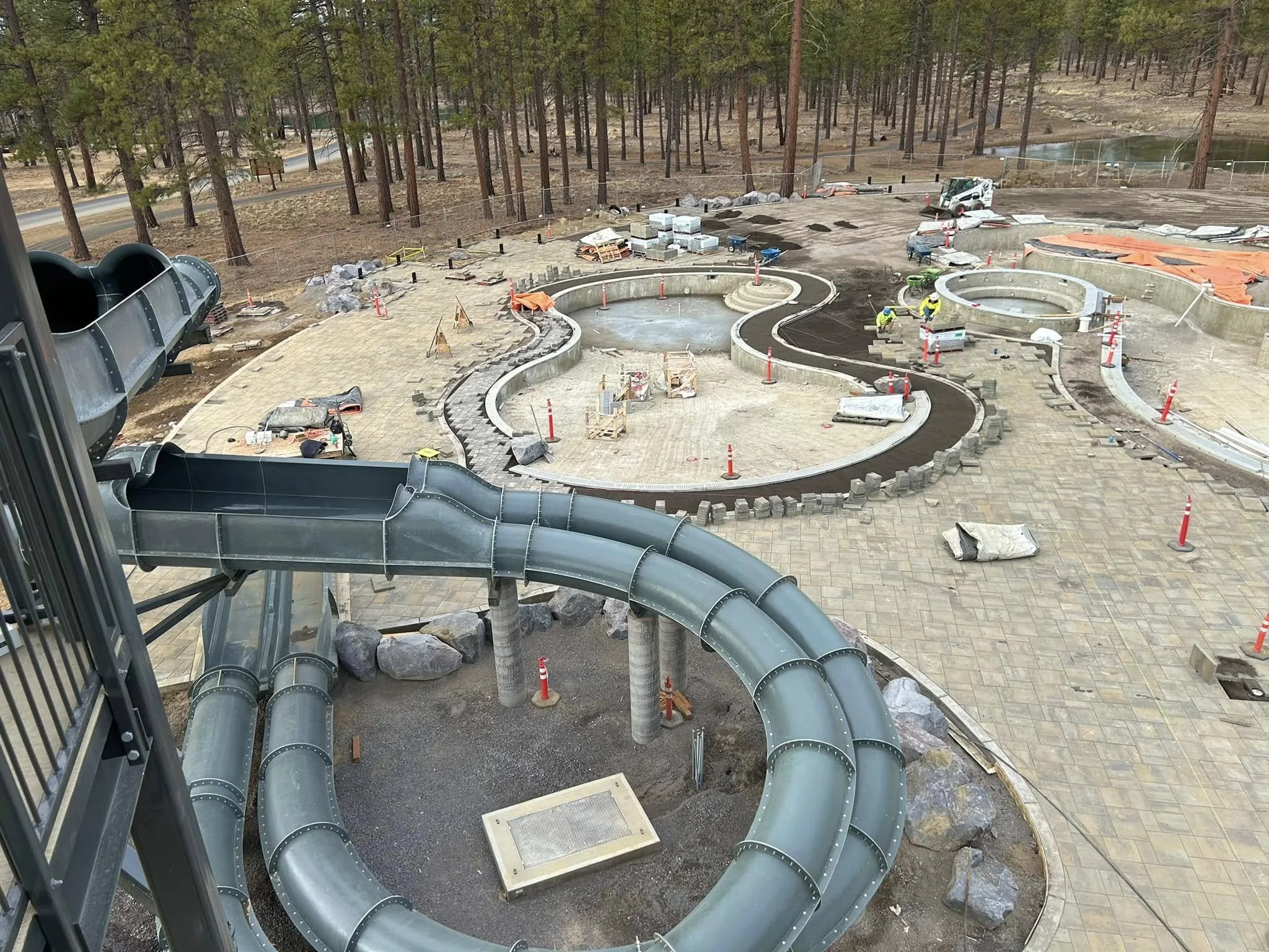 Construction site for a new water park with multiple water slides, some still under construction, surrounded by trees and with construction workers working on the paved area.