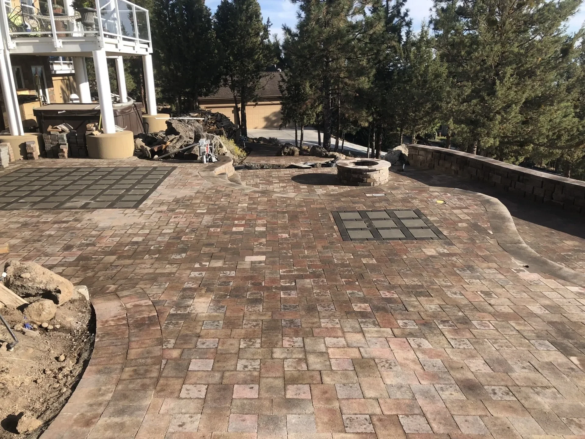A newly paved outdoor patio with brick pavers, a brick fire pit, and a curved stone retaining wall. There are tall pine trees in the background and part of a raised deck with white railings is visible on the left side. The scene is in a natural, wooded setting.