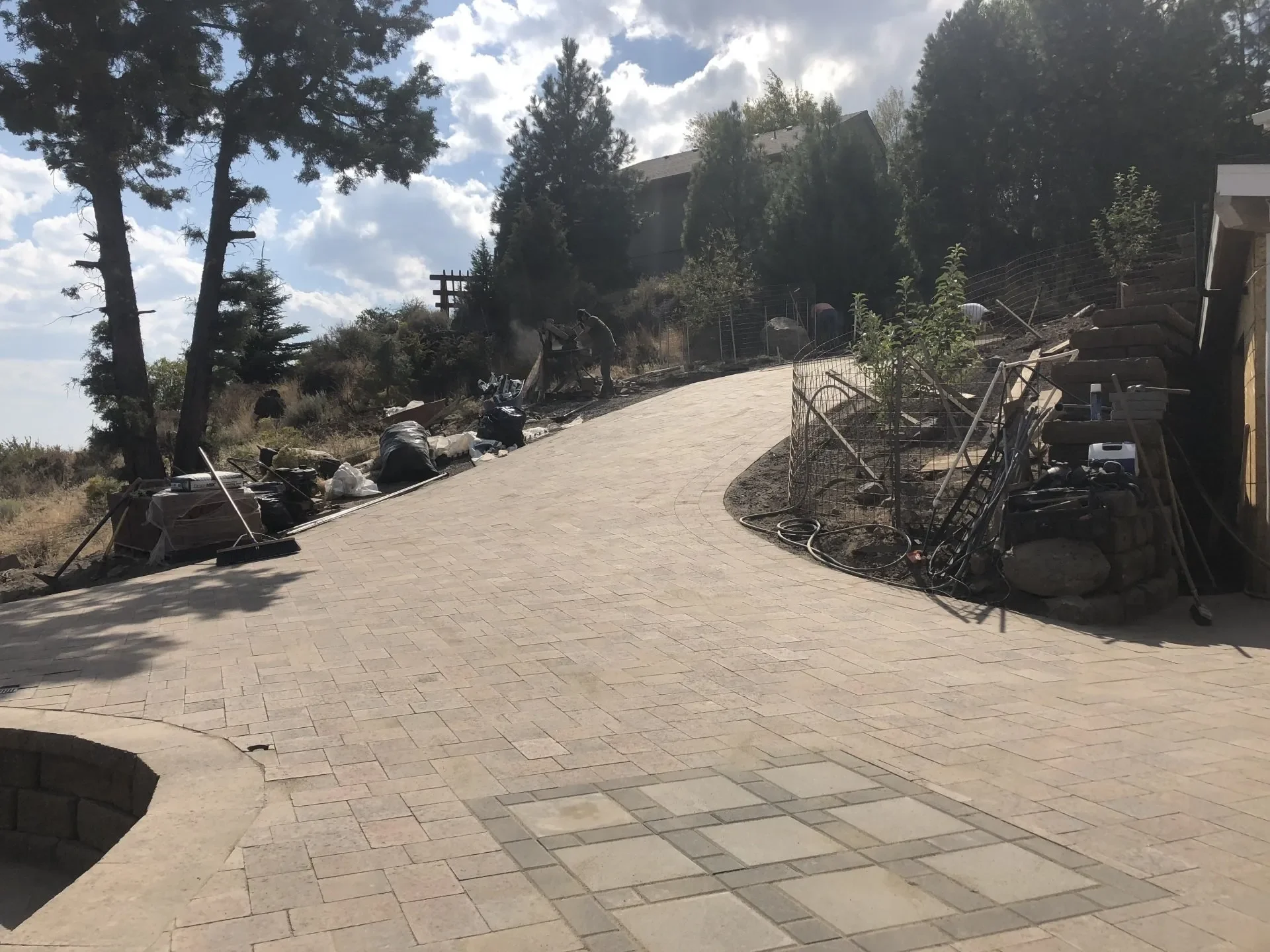 A curved paved pathway on a hillside with trees and a house in the background. There are cluttered objects, tools, and equipment along the side of the pathway, indicating landscaping or construction work.