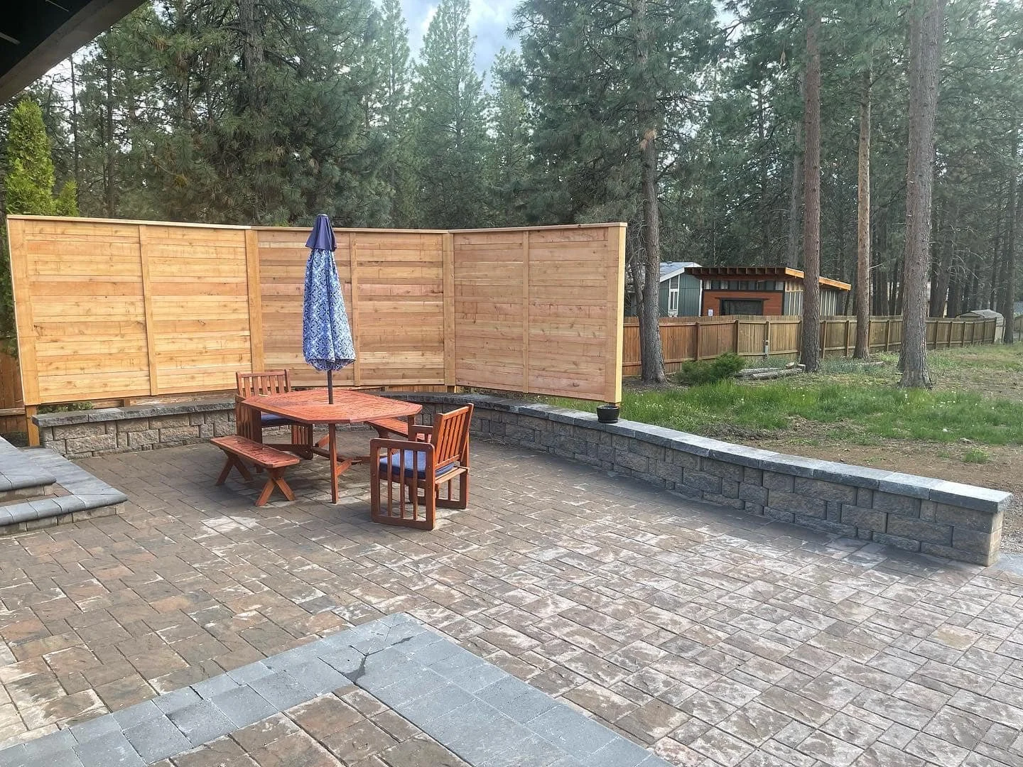Backyard patio with a wooden dining table, benches, chairs, and a large blue umbrella, surrounded by a wooden privacy fence, with trees and a small shed in the background.