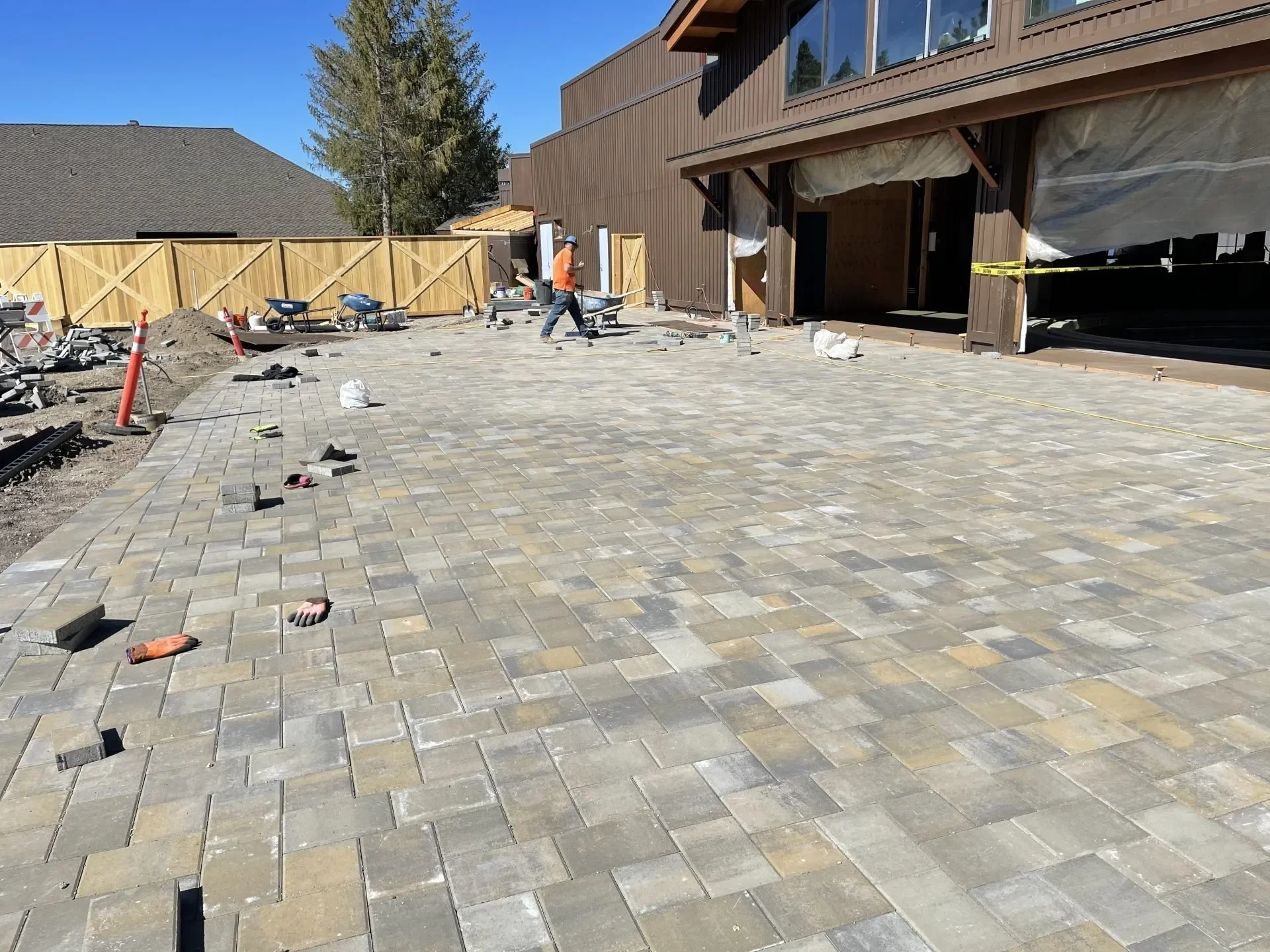 Construction workers installing paving stones in the backyard of a house, with a wooden fence along the perimeter and a building exterior in the background.