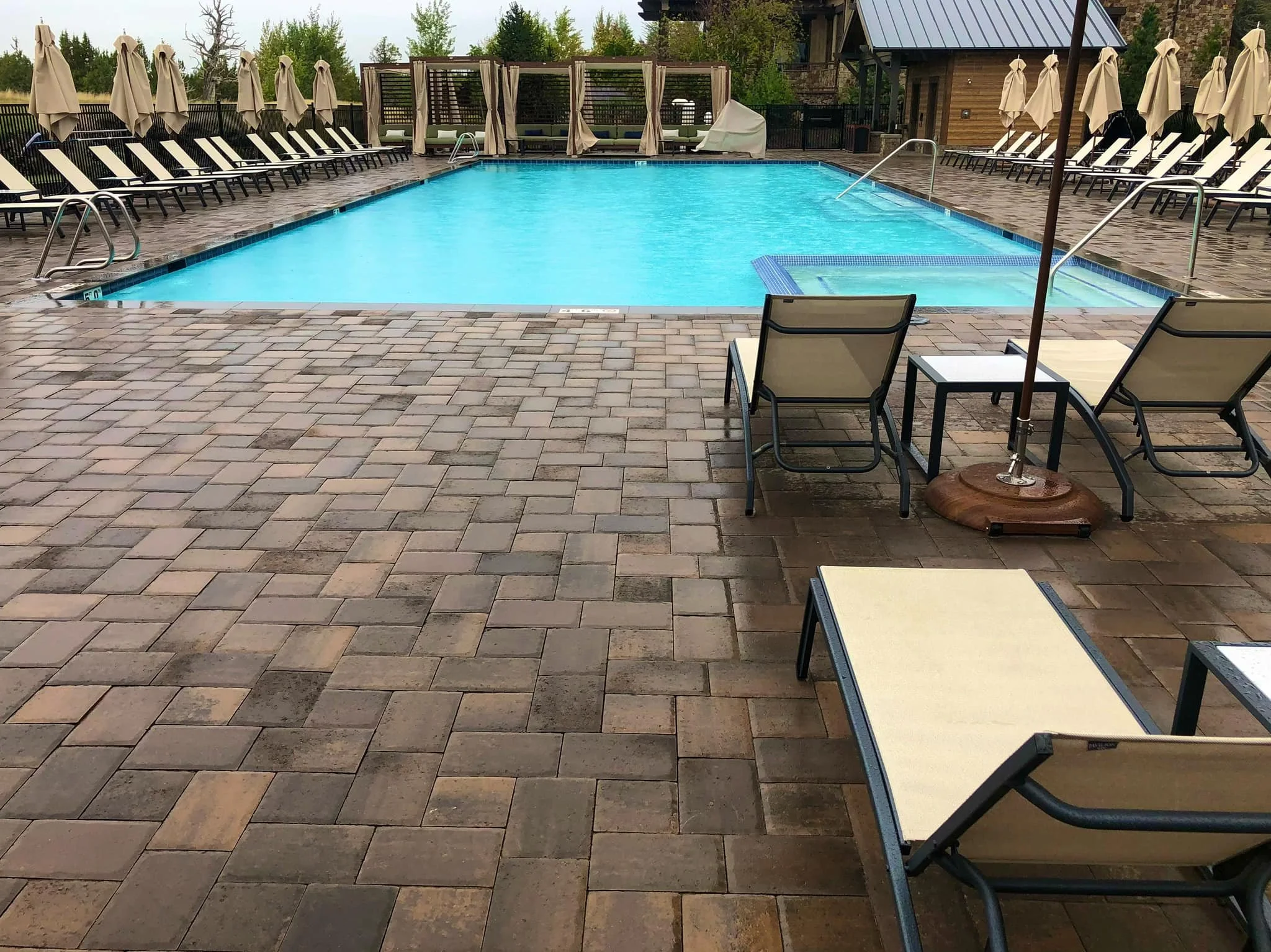 An outdoor swimming pool area with lounge chairs under umbrellas, a shaded cabana, and a brick patio, surrounded by trees and a wooden building.