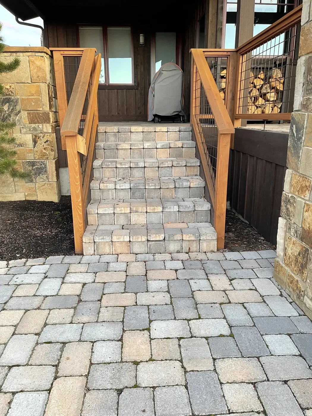 View of a front porch with brick stairs leading up to a wooden entrance, stone and brick walkway in the foreground, and stacks of firewood on the side.