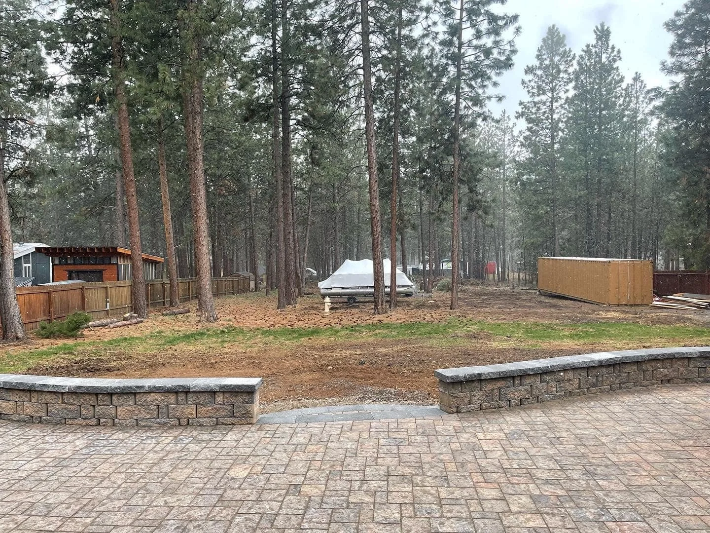 Backyard area with a brick patio, a low stone wall, a grassy patch, tall pine trees, a white covered boat, and construction materials including a wooden fence panel and a pile of wood in the background.