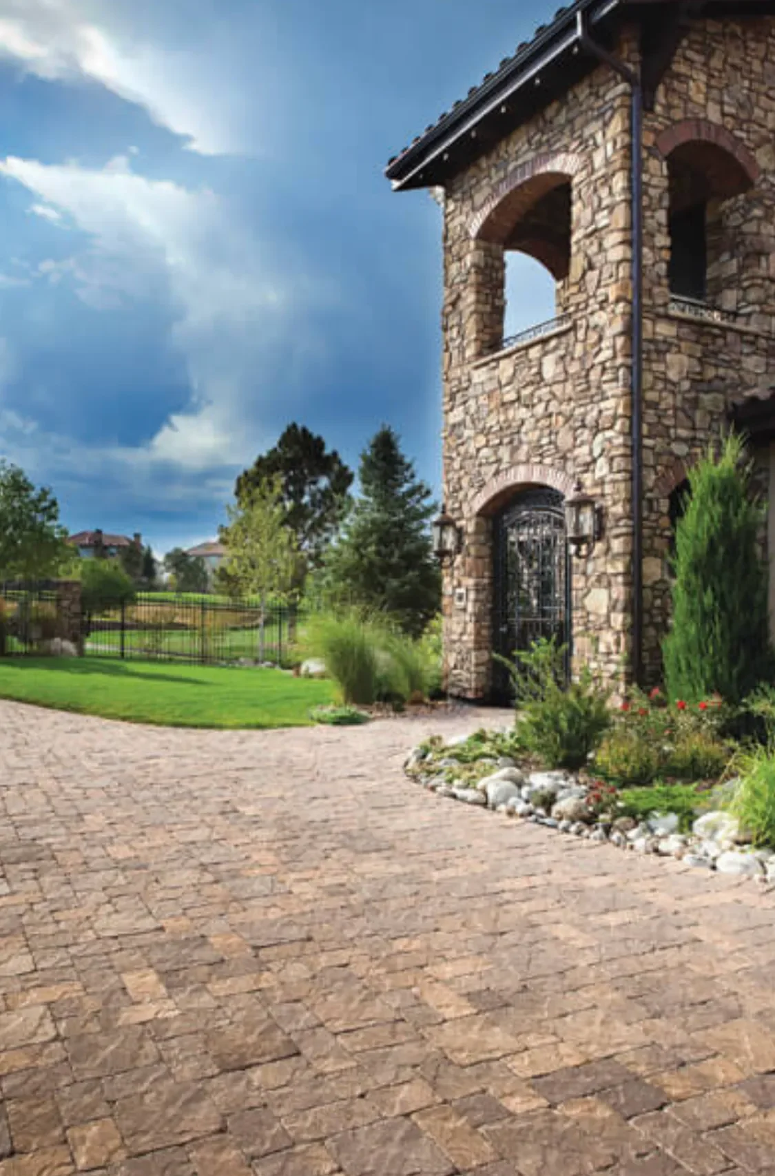 Stone house with arched windows and gated entrance, surrounded by landscaping and a paved driveway, under a partly cloudy sky.