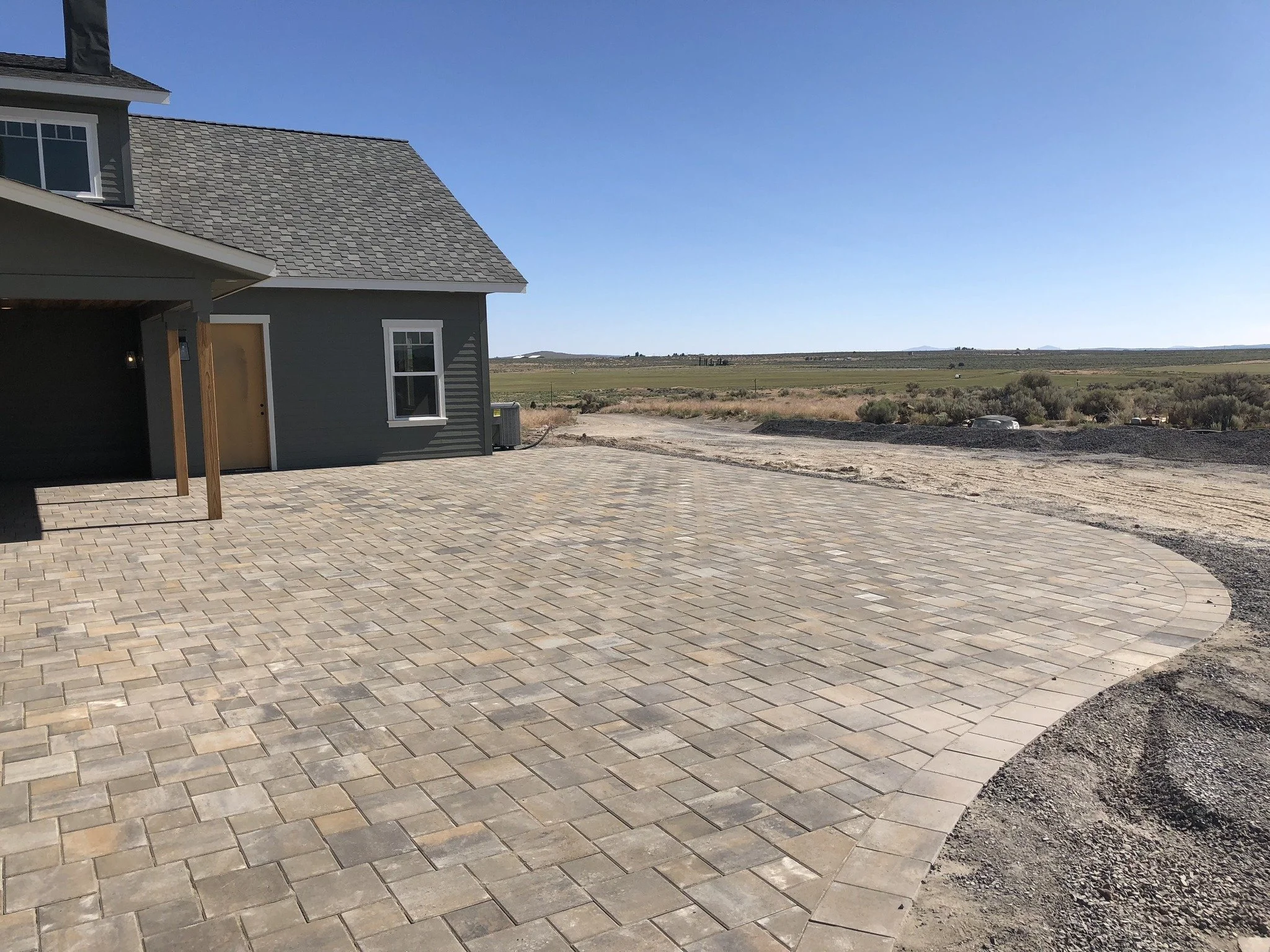 A house with a gray exterior and a tiled roof under a clear blue sky, with a paved driveway and open fields in the background.