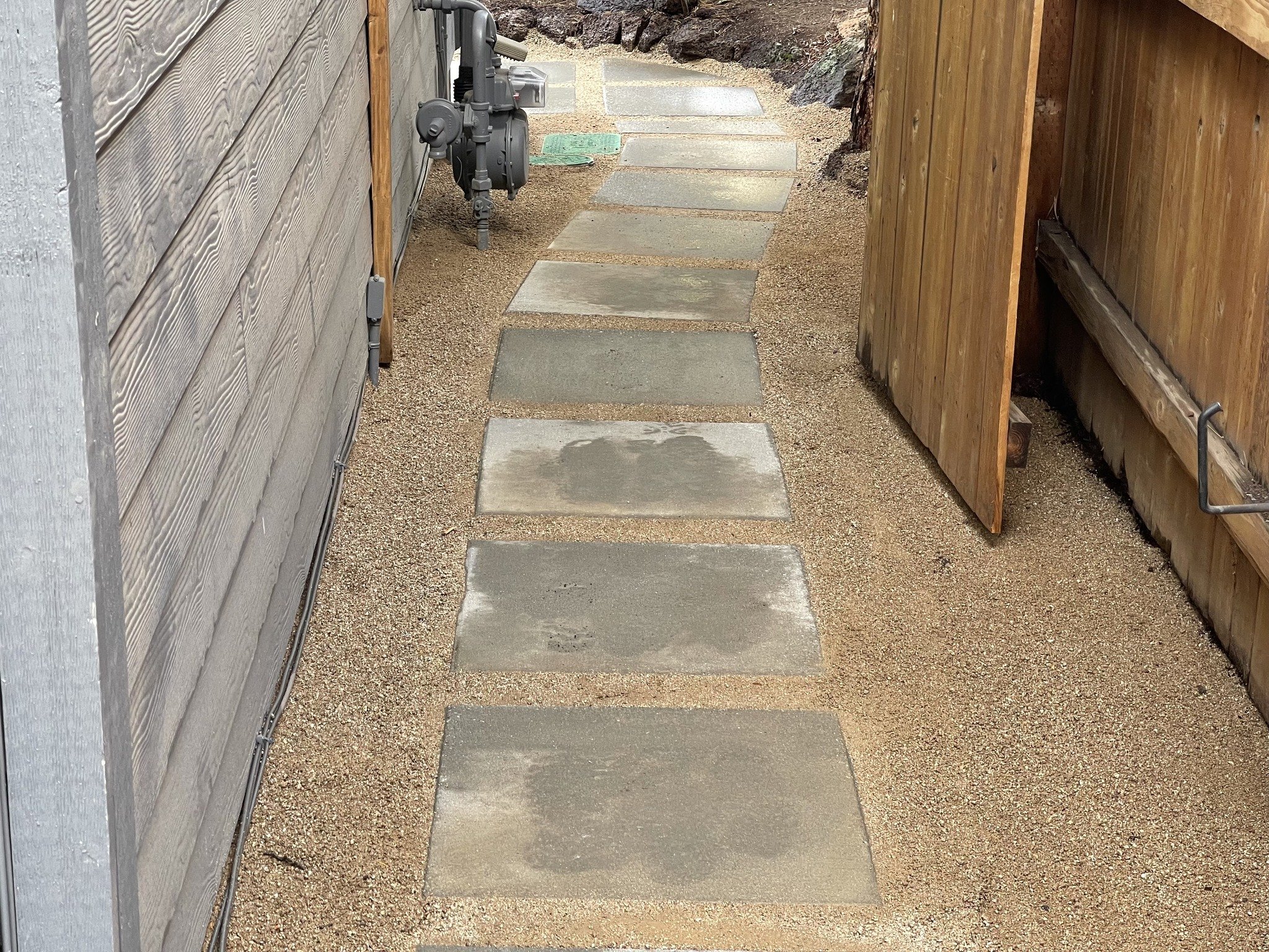 A narrow outdoor pathway with evenly spaced concrete stepping stones, flanked by a wooden fence on the right and a house wall on the left. Gravel surrounds the stones, and there is outdoor equipment attached to the house wall.