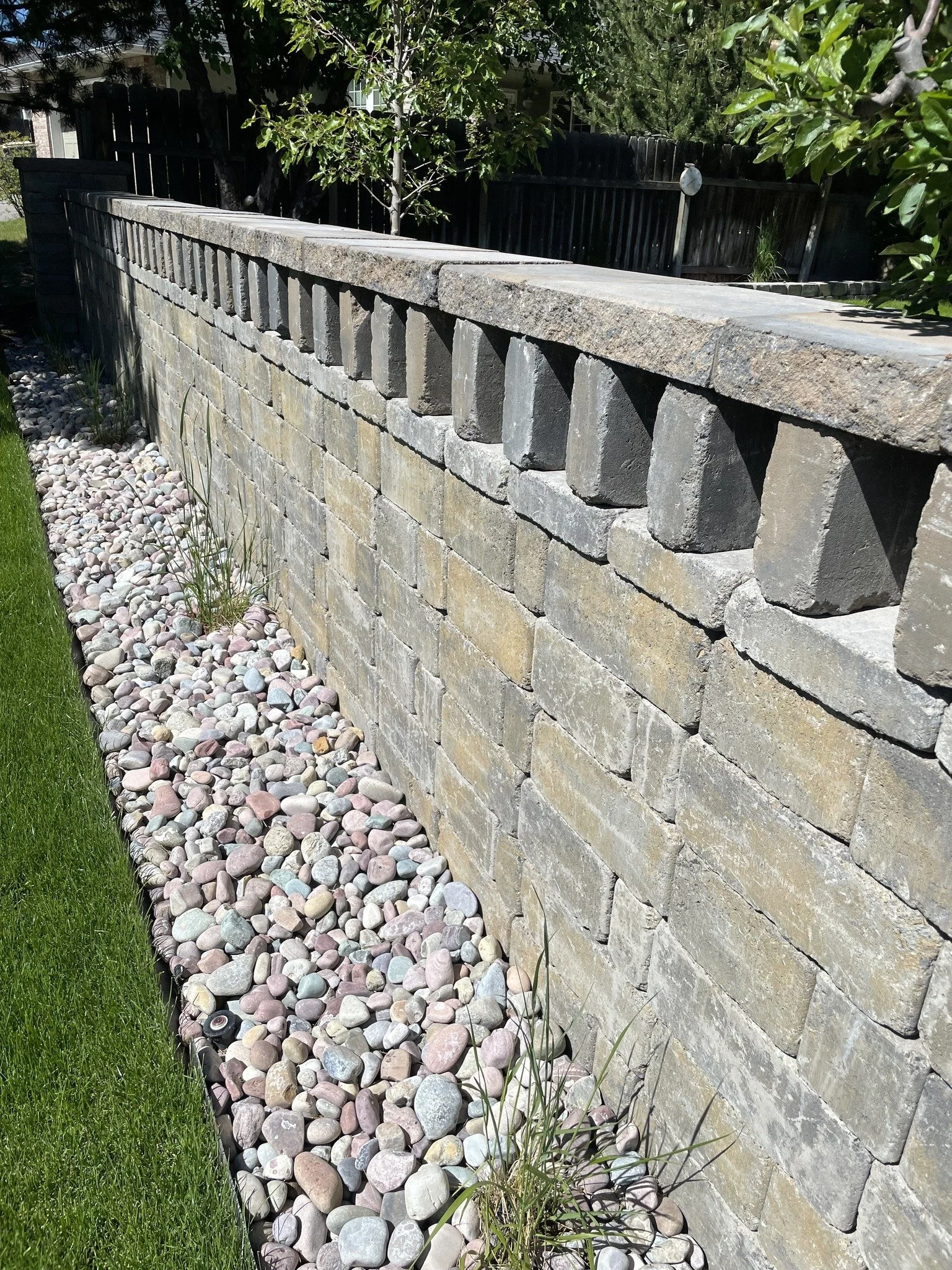 A stone retaining wall with a bed of multicolored smooth river rocks at its base, surrounded by green grass and trees.
