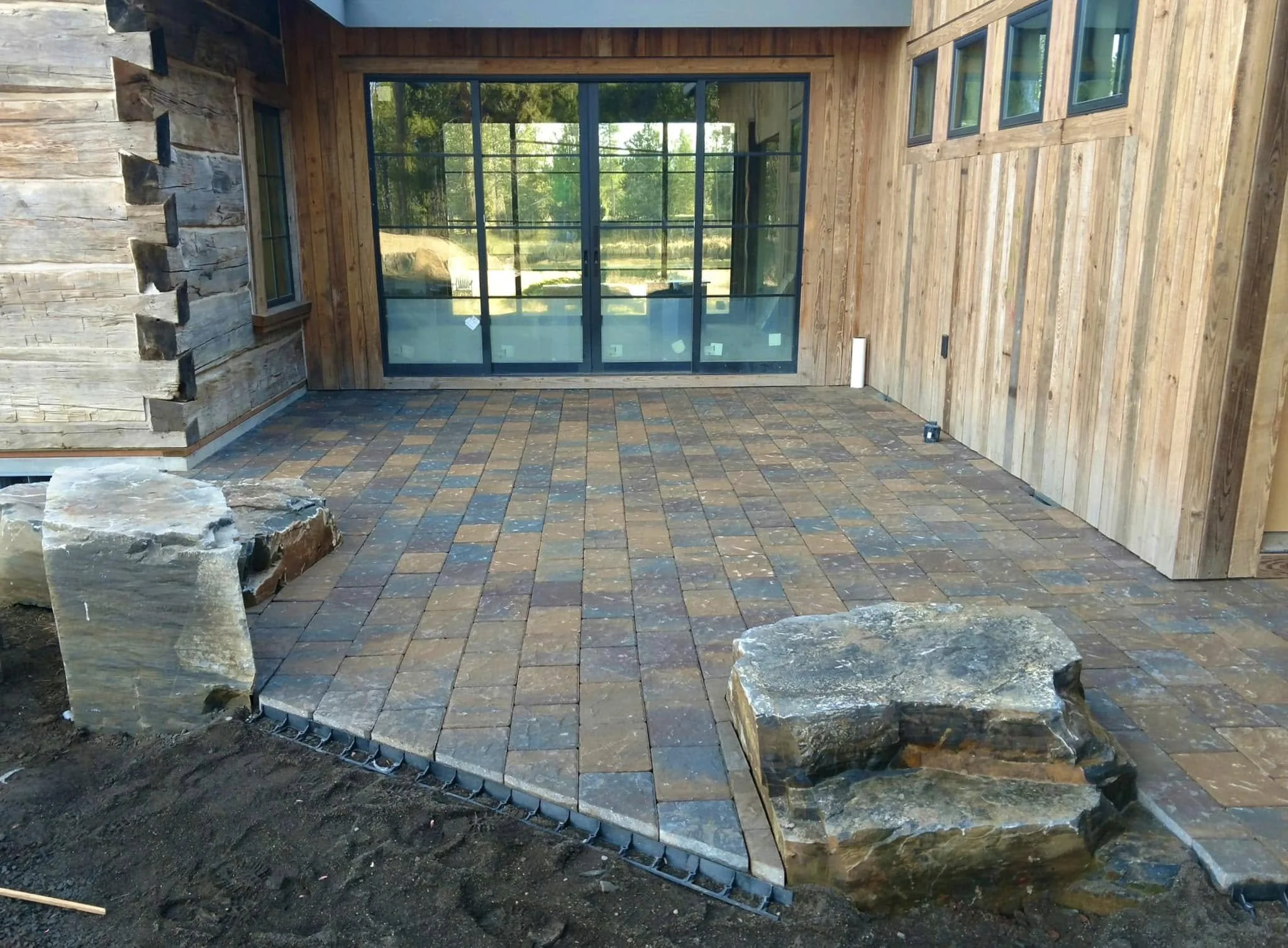 A backyard patio with brick pavers, partly enclosed by wooden walls and large glass sliding doors, with rocks placed along the edge.