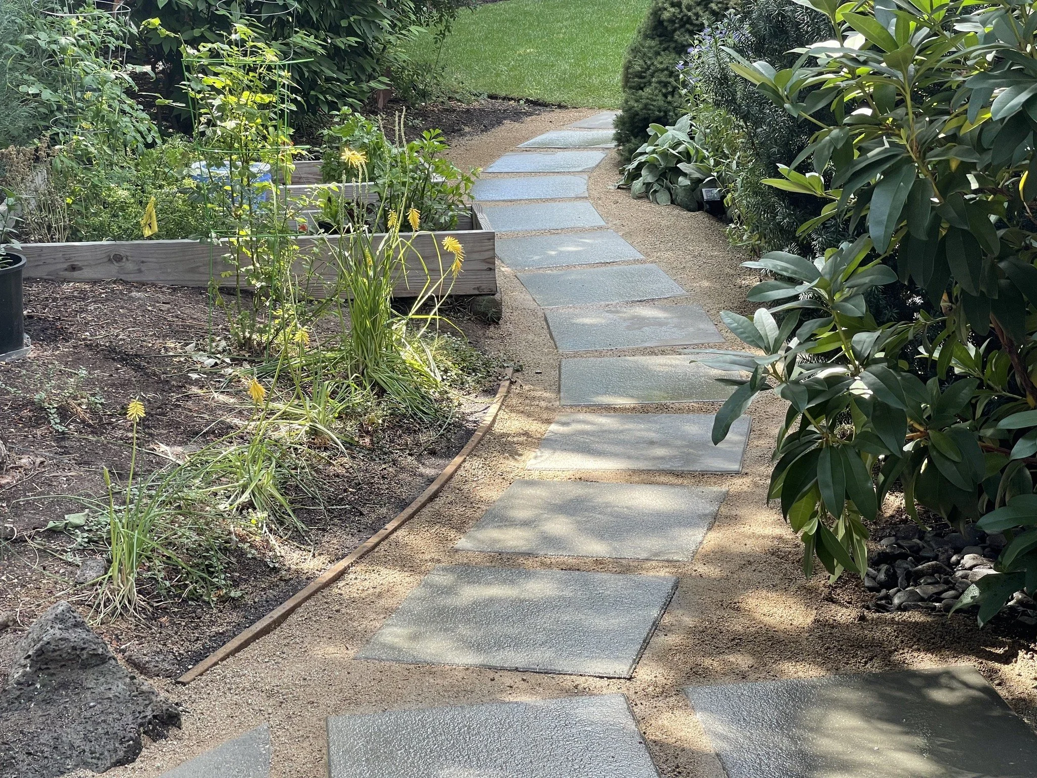 A garden pathway with rectangular stone slabs bordered by soil and greenery on both sides, including bushes, small plants, and grass.