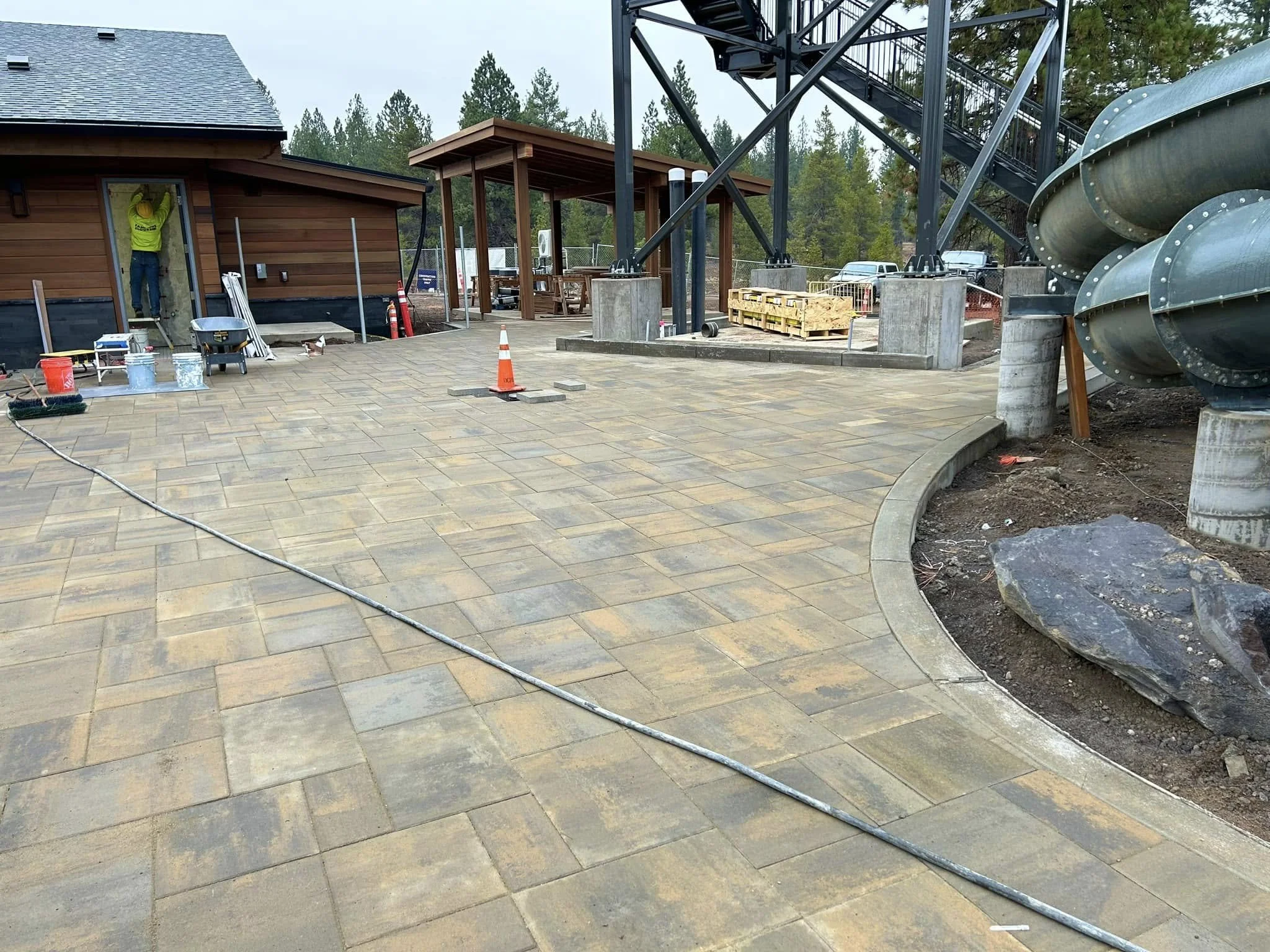Construction site with workers installing new pavement, a small wooden building, and a large metal slide or tube structure in the background surrounded by trees.