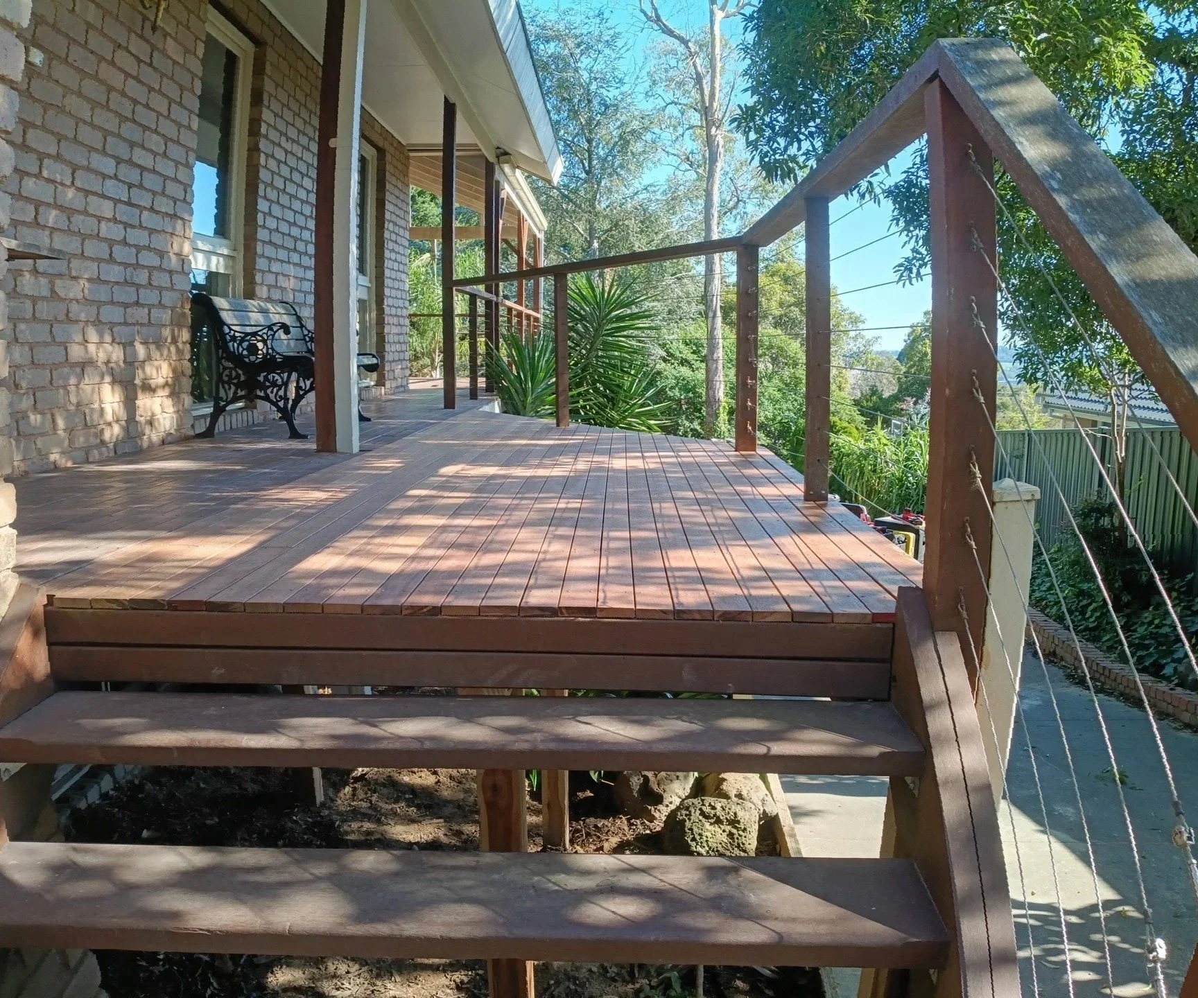 Wooden deck attached to a brick house with a black metal bench, surrounded by greenery and trees, with a view of the sky. elevate property maintenance and repairs home care landscaping melbourne outer east and yarra valley deck build and repair 