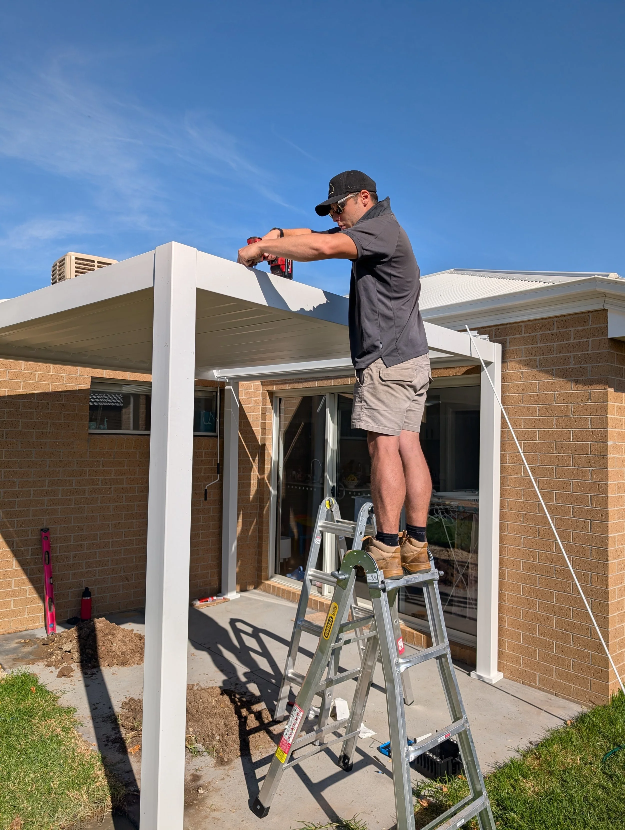 A man wearing a gray shirt, khaki shorts, a black baseball cap, and sunglasses is standing on a step ladder while working on the corner of a white pergola attached to a brick house. elevate property maintenance and repairs home care landscaping 
