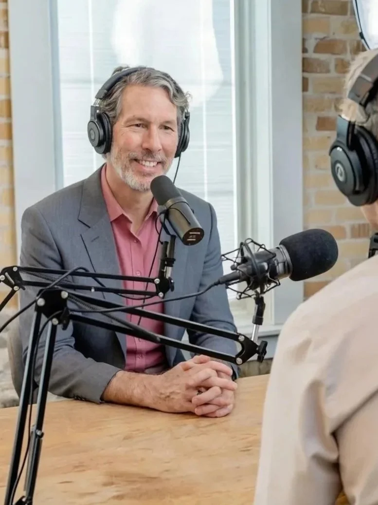 A man with gray hair and a beard, dressed in a gray suit and pink shirt, sitting at a table with a microphone, headphones, and recording equipment, speaking into a microphone during a podcast or radio interview.