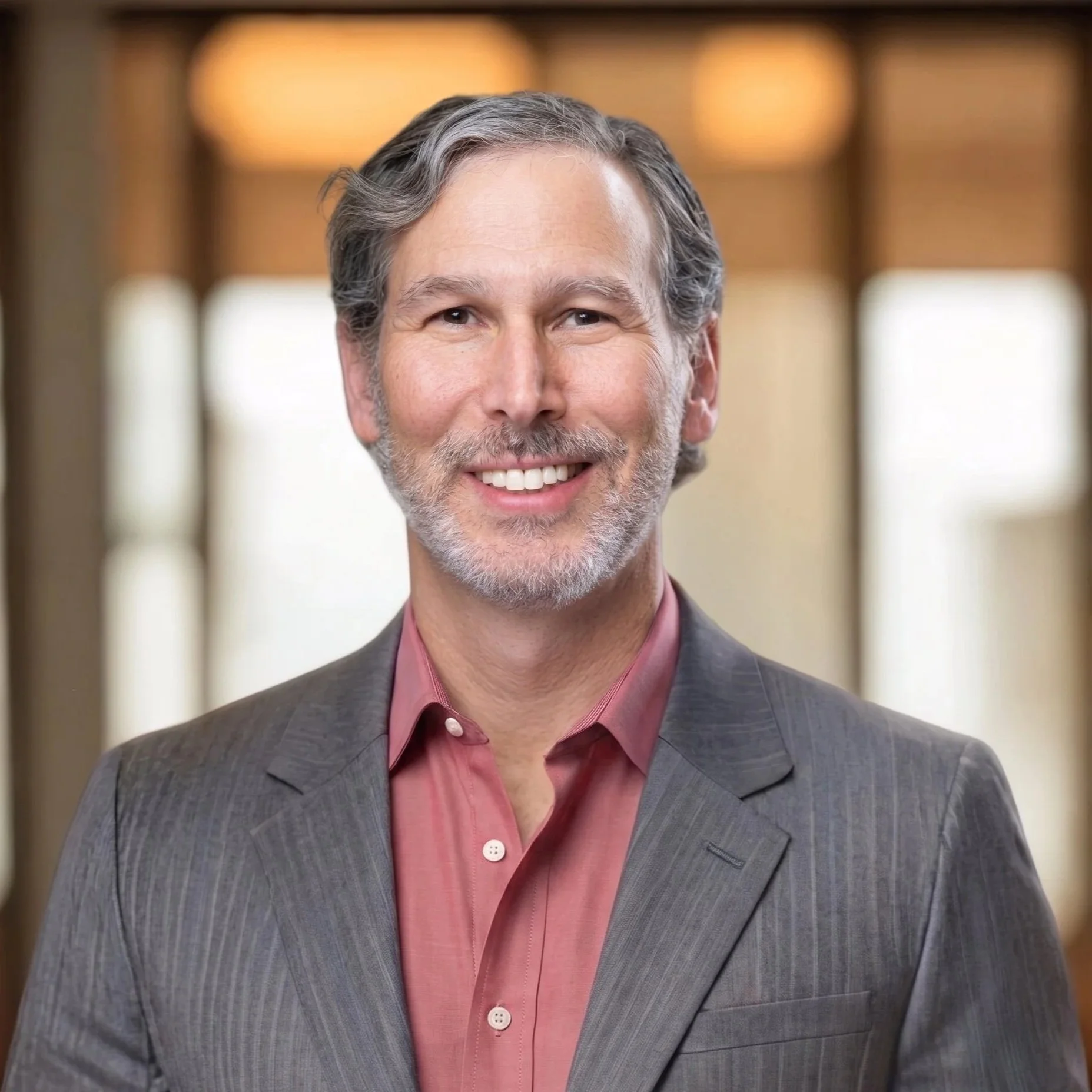Headshot of a smiling middle-aged man with gray hair and beard, wearing a dark gray blazer and a pink button-up shirt, standing in a room with a blurred background.