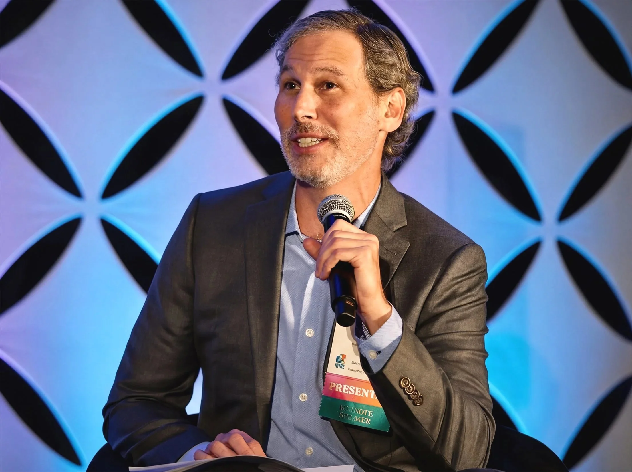 A middle-aged man with gray hair and a beard, wearing a dark suit jacket and light blue shirt, speaking at an event. He is holding a microphone in his right hand. The background features a blue and white patterned backdrop. The man has a pink and green badge hanging from his neck with the words "PRESENT" and "KOUNOTE SPEAKER" visible.