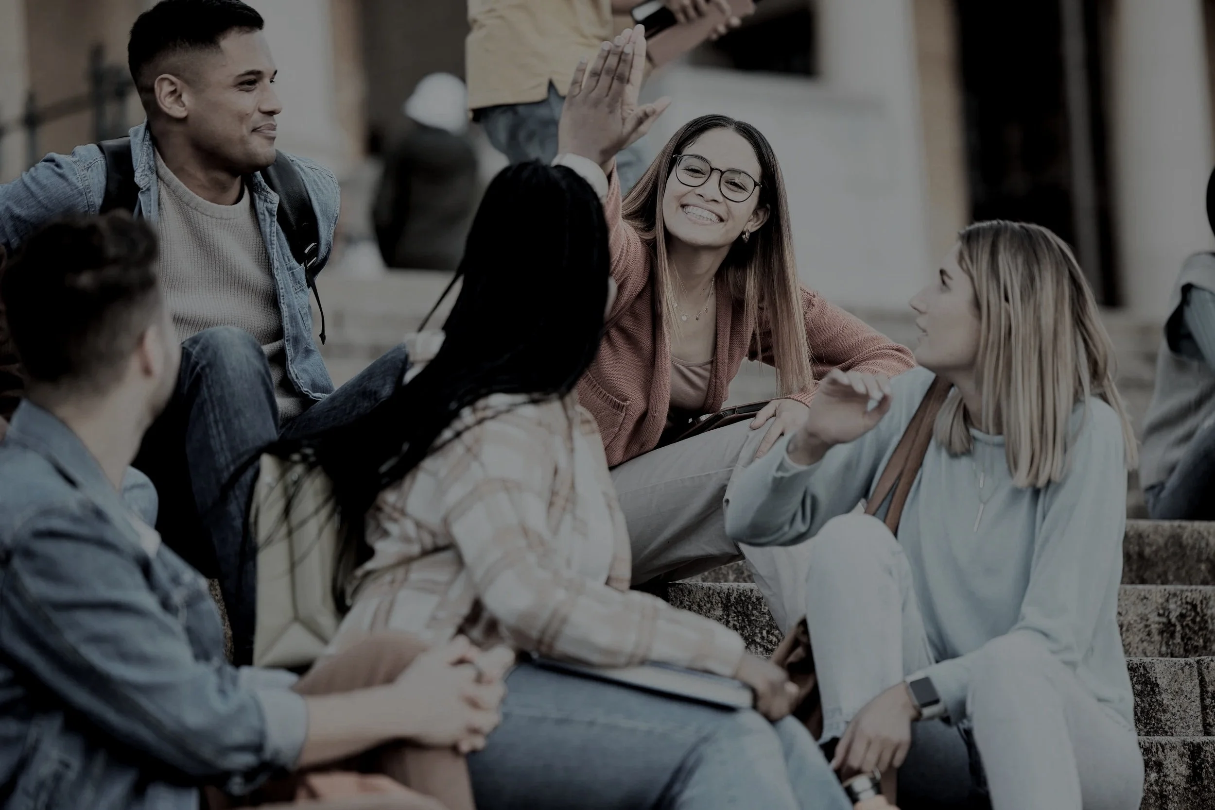 Group of diverse young adults sitting on steps outdoors, smiling, engaging in conversation, and one woman raising her hand for a high-five.