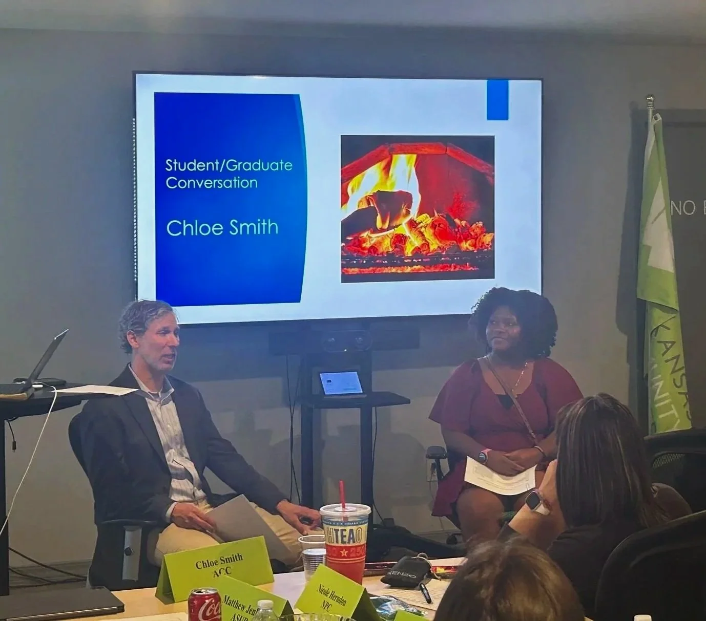 A presentation with a slide on a large screen reading "Student/Graduate Conversation, Chloe Smith," accompanied by an image of a campfire. Two women are seated and speaking, with one holding a paper and the other with a notebook. A table with name tags, drinks, and electronic devices is in the foreground.
