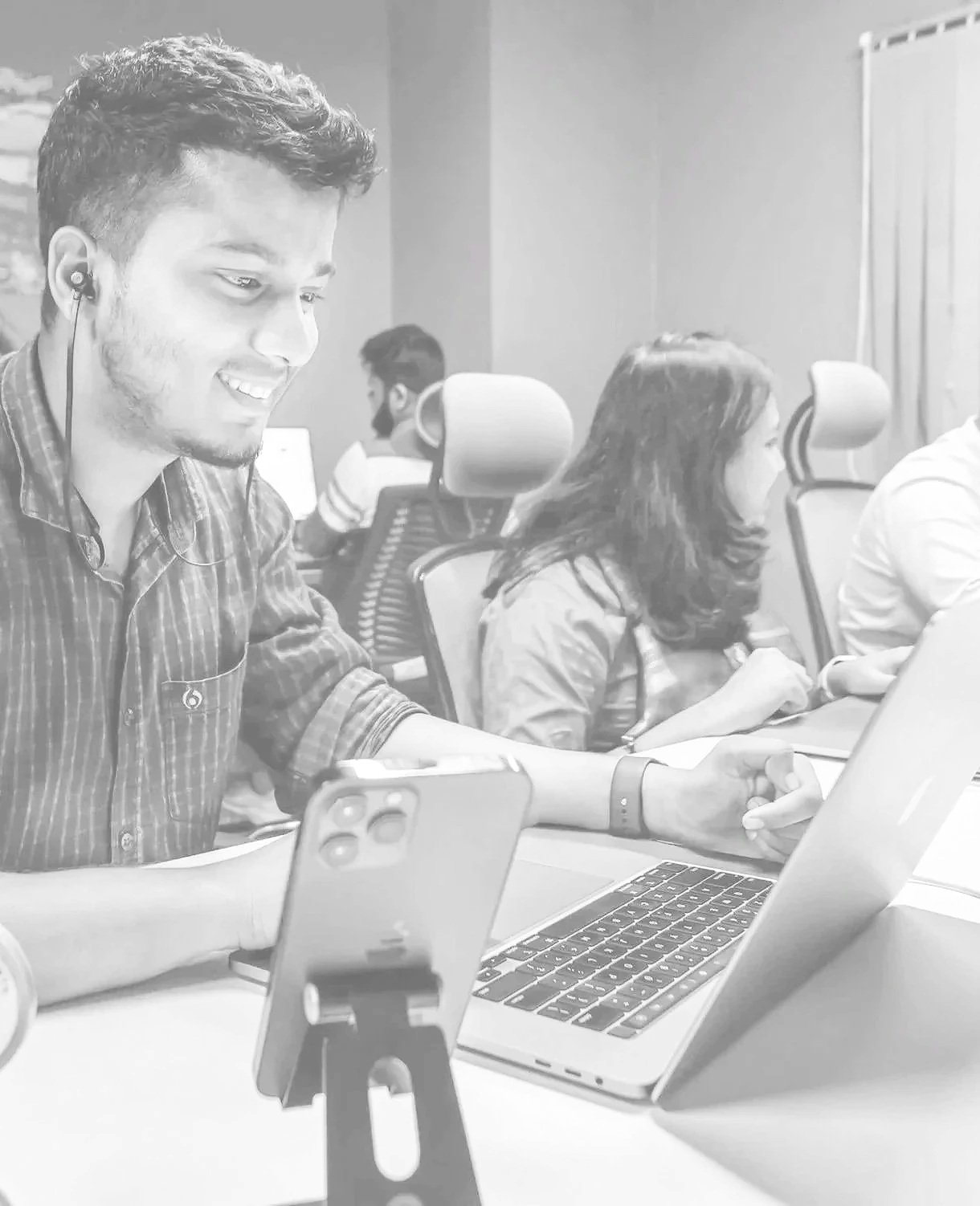 Young man with earbuds smiling at his phone, working on a laptop in an office with others in the background.