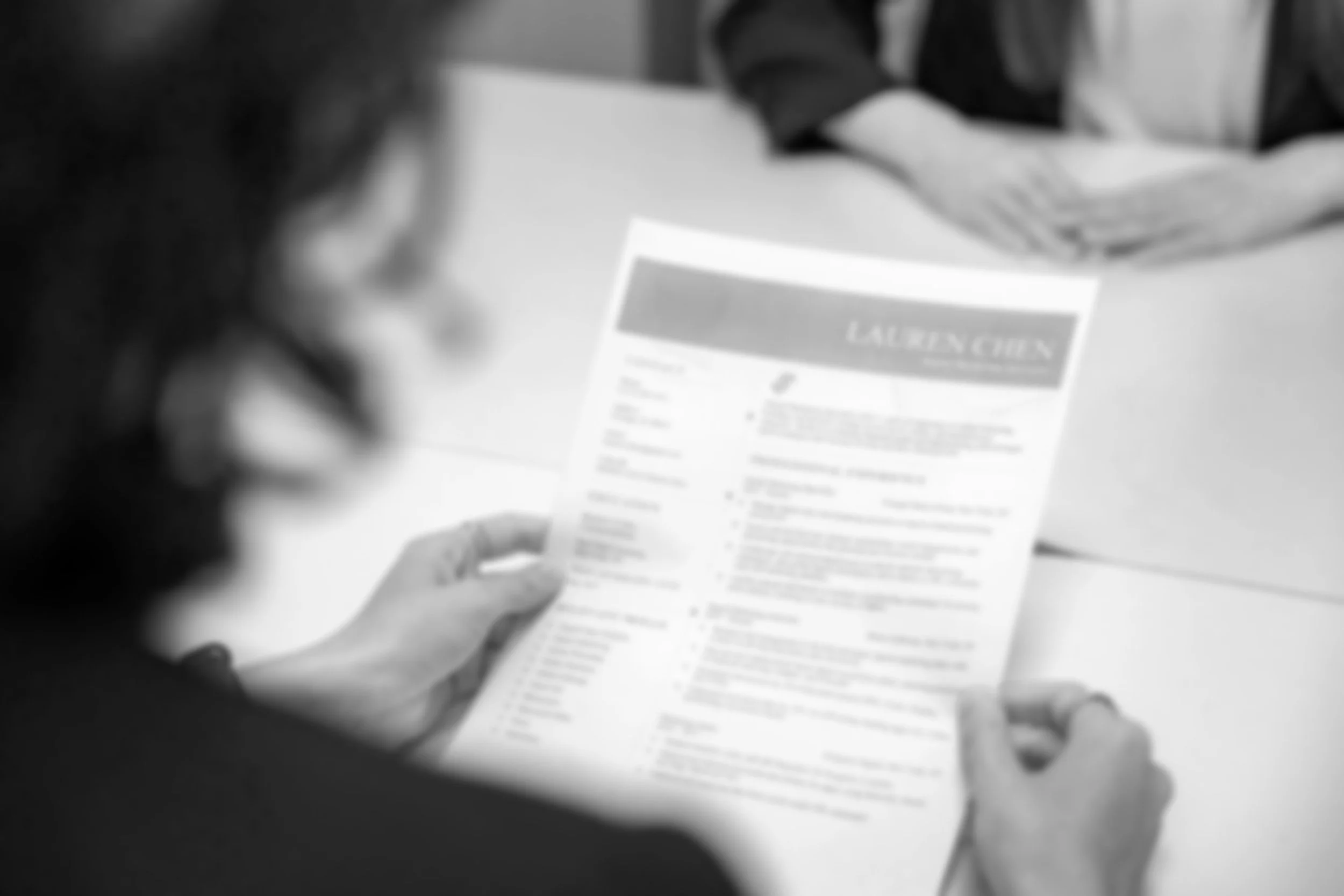A person's hand holding and looking at a printed resume or document, with the word 'LAUREN CHU' visible at the top. The background shows a table or desk.
