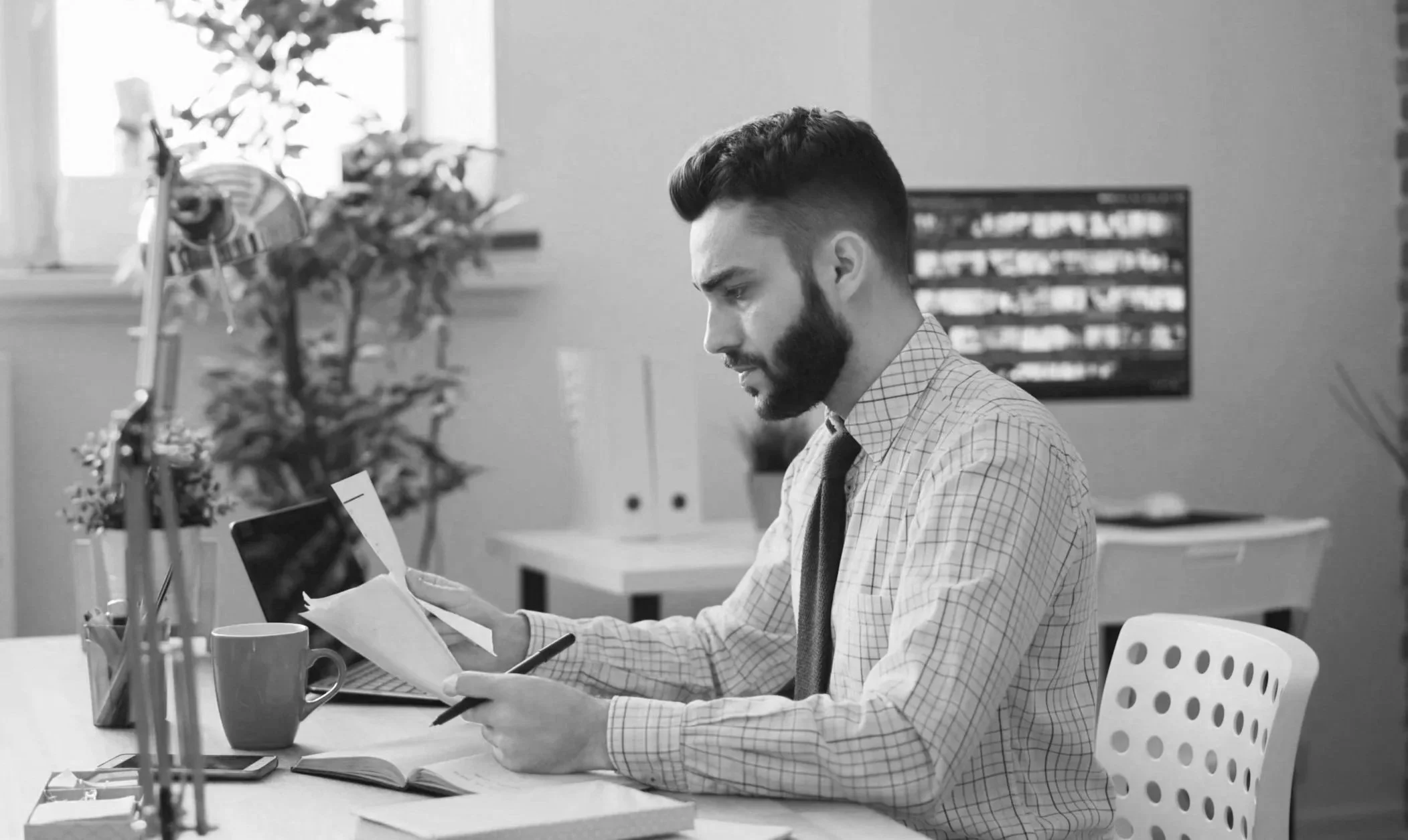 A man in a checkered shirt and tie sitting at a desk, looking at a tablet or phone. There are a computer and coffee cup on the desk, with a window and some plants in the background.