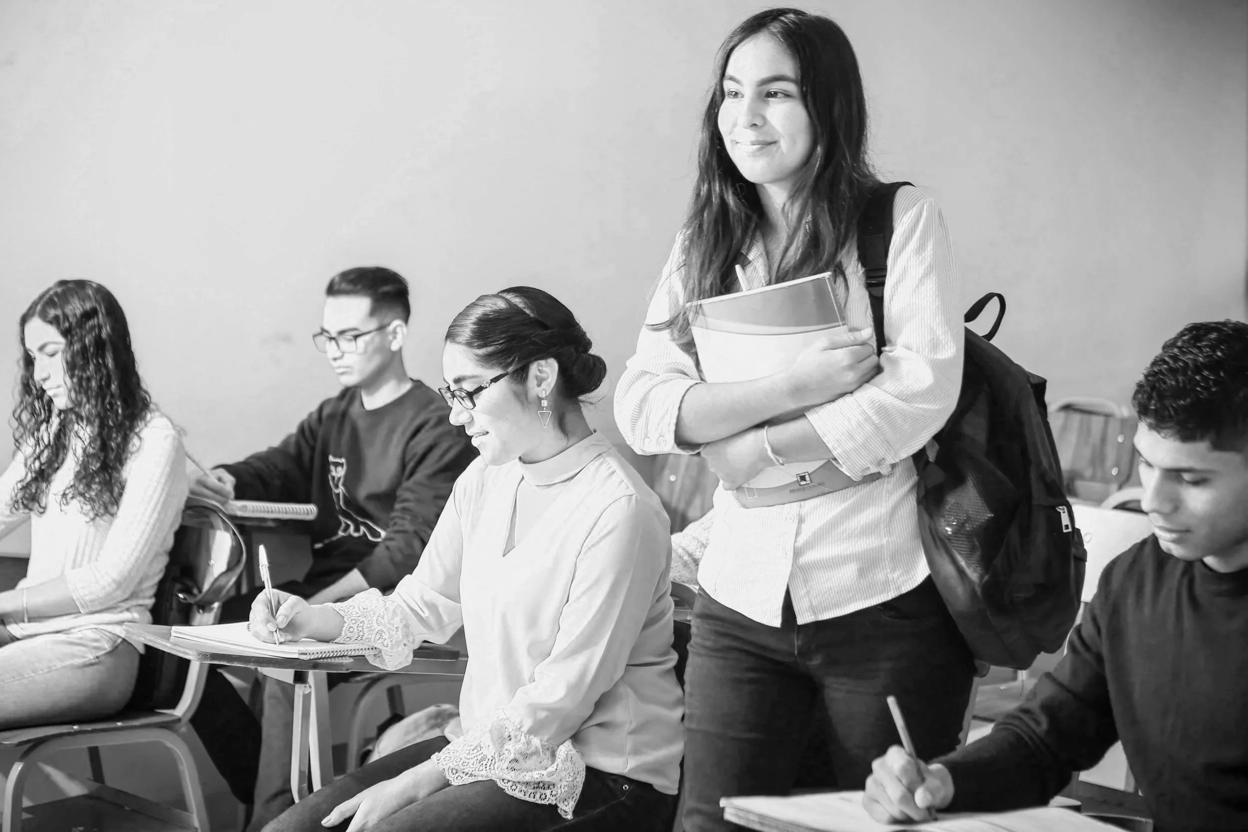 Students sitting at desks in a classroom, with one standing student holding books and looking to the side.